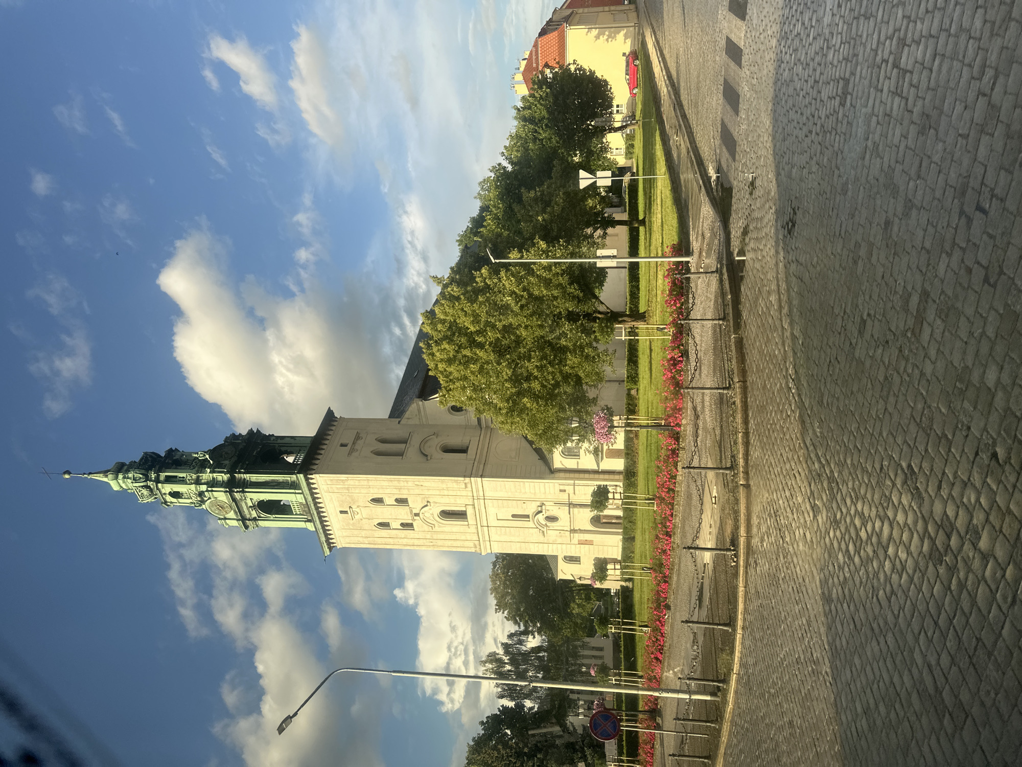 The image shows the Church of the Exaltation of the Holy Cross in Krosno Odrzańskie, Poland. The church is a prominent landmark, with a tall, pale stone tower topped by a green copper spire. The tower has several levels, including arched windows and a clock face. The main body of the church is visible to the right of the tower and appears to be constructed from similar stone. The church is surrounded by mature green trees and a well-maintained lawn adorned with a border of pink flowers. In the foreground, a cobblestone street curves around the church grounds, with a pedestrian crossing visible. A street lamp stands on the left side of the street, and a red car is parked in front of a building with a red-tiled roof on the right side of the image. The sky is blue with scattered white clouds, indicating it is daytime. The scene appears peaceful and well-kept.