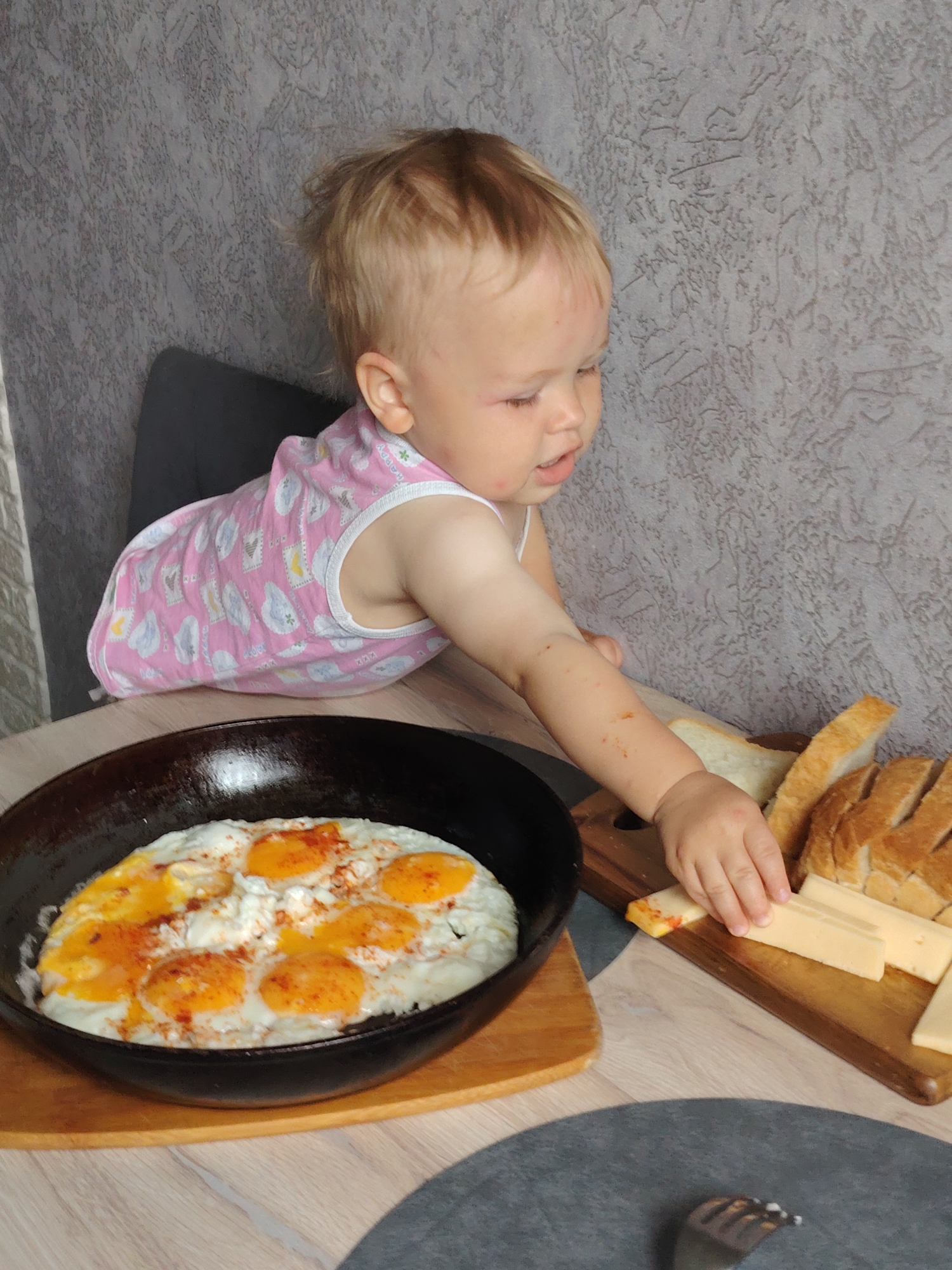A young child is reaching for a slice of cheese on a wooden cutting board. The cutting board also holds sliced bread. A frying pan with several fried eggs sits on a wooden surface next to the cutting board. The child is wearing a pink tank top. The location is Chaikovsky, Russia, based on the information provided.
