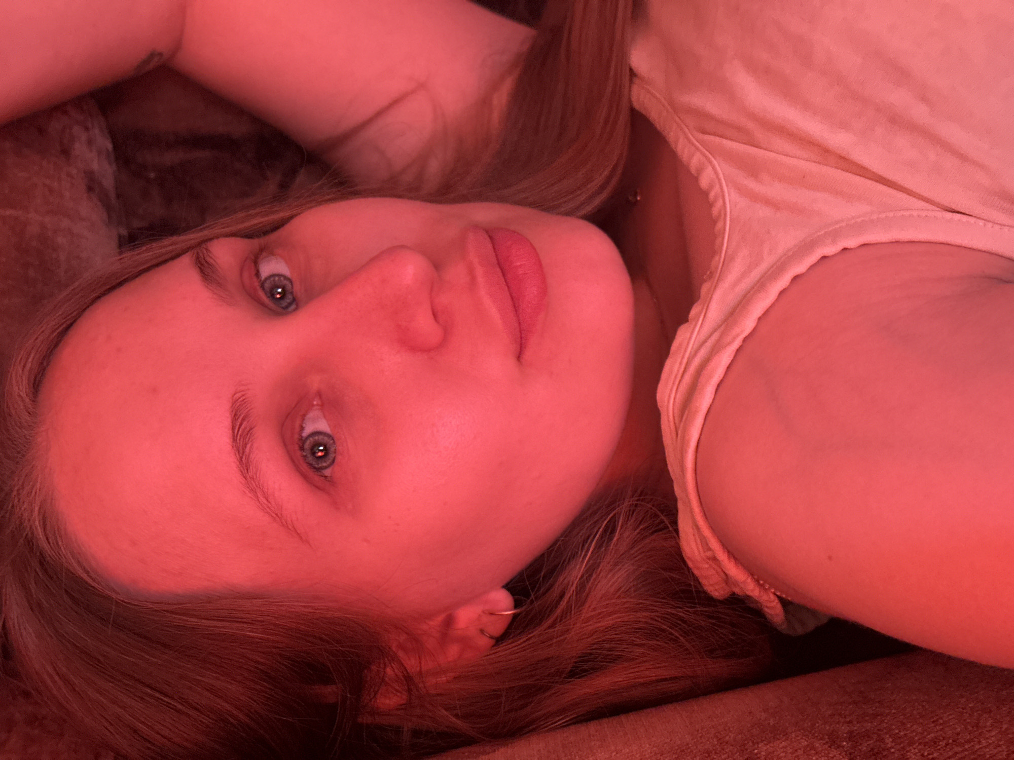 A fair-skinned person with light eyes and reddish-brown hair is lying down. The person is wearing a light-colored shirt. The scene is bathed in red light. The location is unspecified.