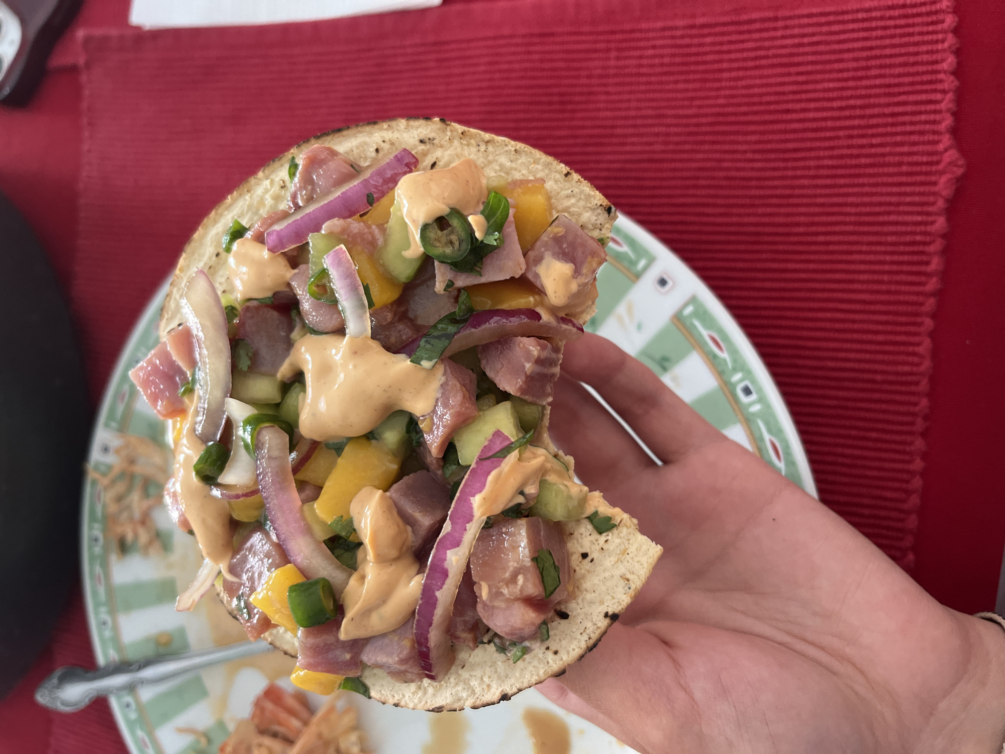 The image depicts a person holding a tostada topped with what appears to be tuna poke. The poke consists of diced raw tuna, red onion slivers, yellow bell peppers, cucumbers, green onions, cilantro, and a creamy sauce, likely spicy mayo or a similar dressing. The tostada is on a plate with a green and white decorative rim, with a red textured surface in the background. A fork and remnants of other food items are visible on the plate. There is no evidence to ascertain that the image was taken in Barrio de Nuevo León, Mexico.