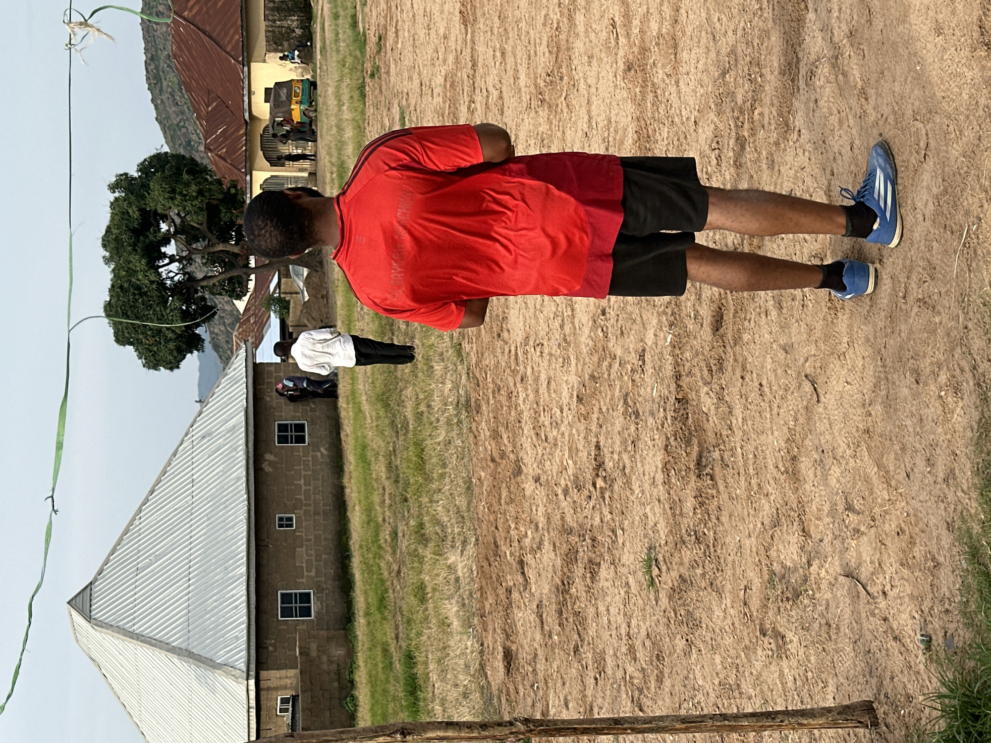An outdoor medium shot captures a man in a red t-shirt and black shorts from the back, standing on a dirt and dry grass field. He is wearing blue athletic shoes and black socks. In the background, a large building with a corrugated metal pyramid-shaped roof is visible, constructed from brown bricks. Several people are gathered near this building. To the right of the main building, another structure with a reddish-brown metal roof is present, and a yellow and green auto-rickshaw is parked in front of it, with several individuals standing nearby. Further in the distance, rolling hills are visible under a pale sky. The scene appears to be in a rural or semi-urban setting in Rukuba Road, Nigeria.