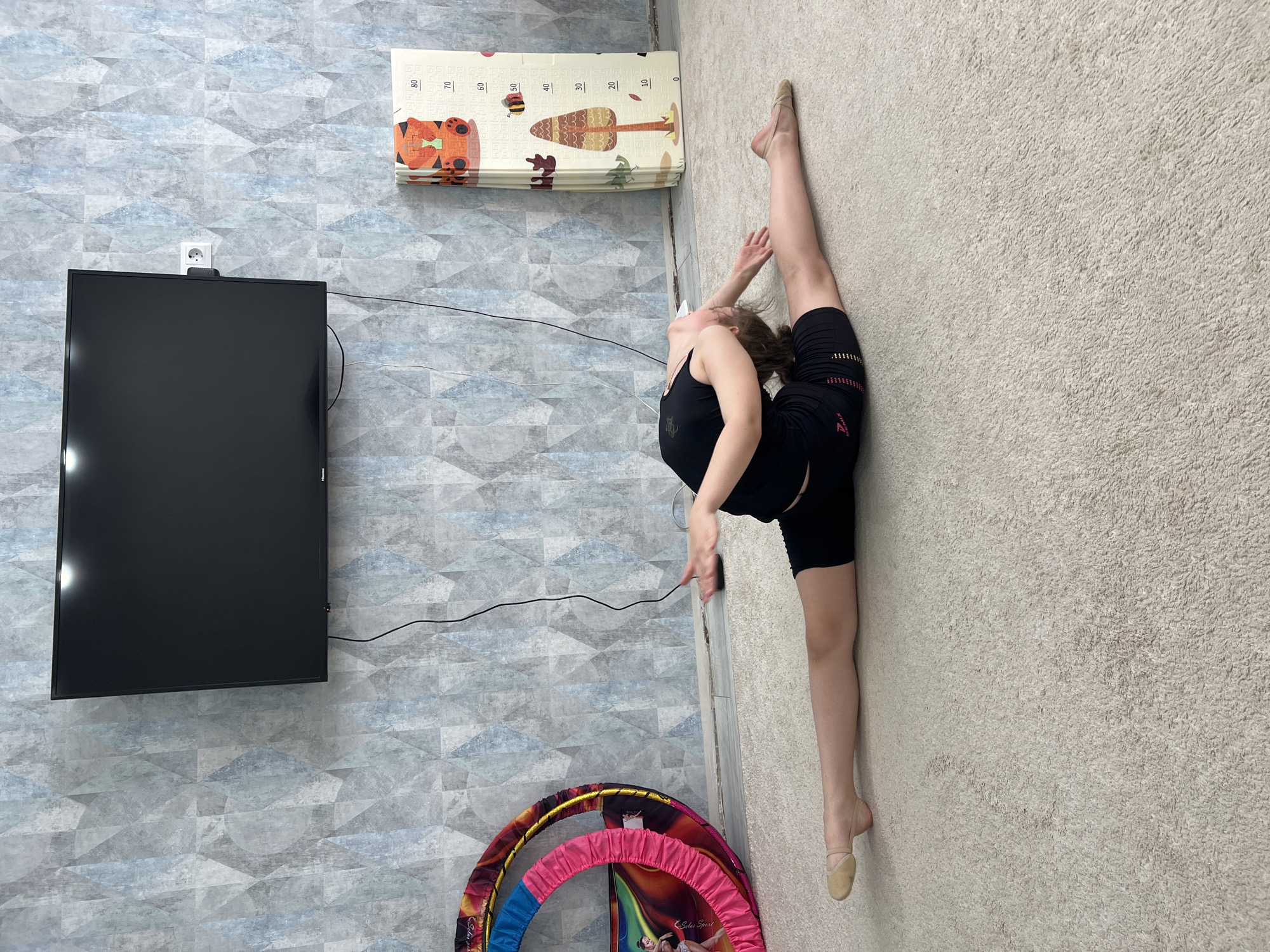 The image shows a woman in a black tank top and black shorts performing a split on a light-colored carpet. Her left leg is extended upward along a wall, while her right leg extends along the floor. She is wearing ballet slippers. Her left hand is arched over her head, while her right arm is extended. A television is mounted on a blue patterned wall to the left, with a power outlet above it. Above the television is a children's playmat. A gymnastics hoop rests near the bottom of the frame. This photo was likely taken inside a home located in the city of Chaikovsky, Russia.