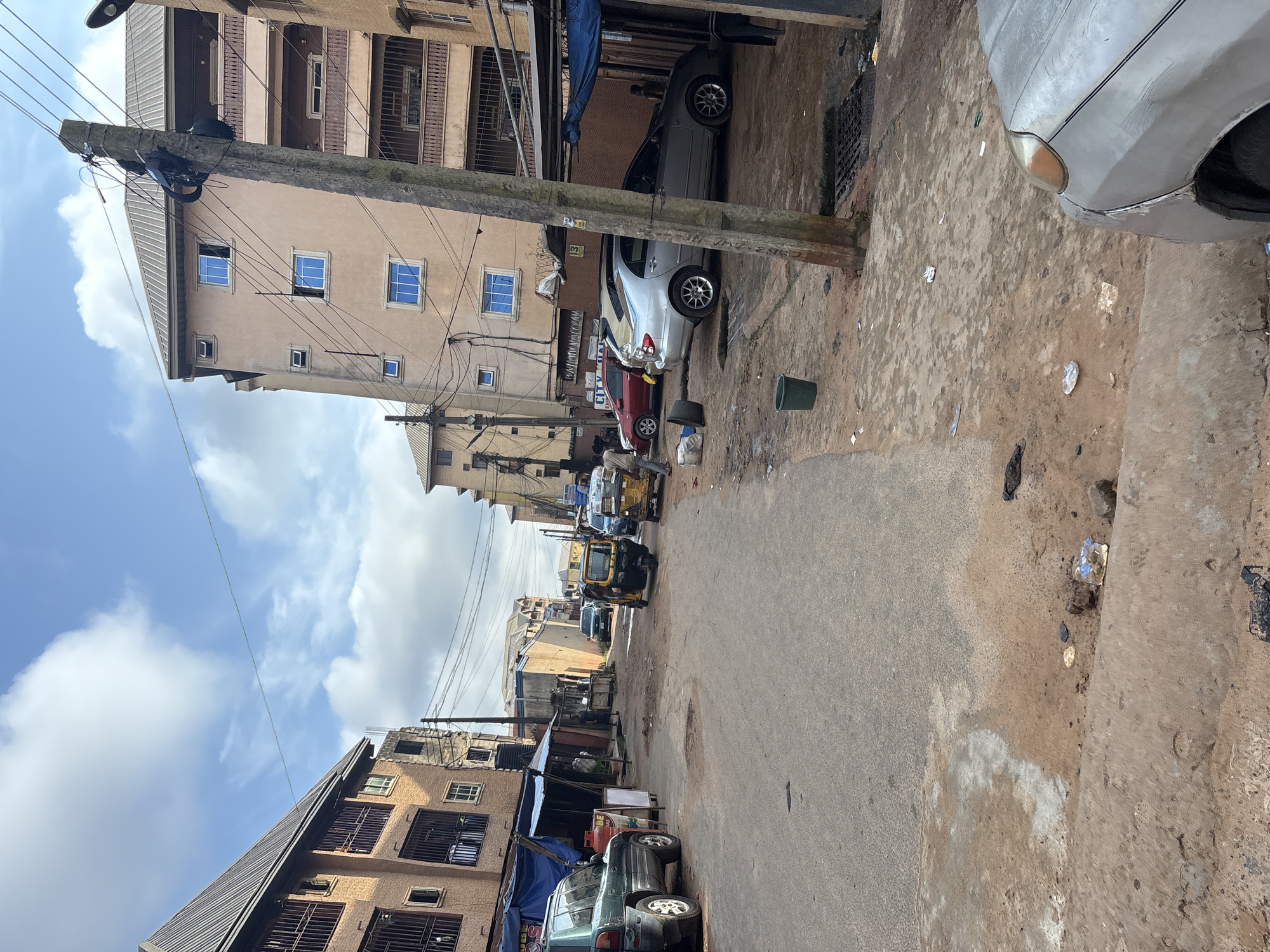 The image depicts a street scene in Onitsha, Nigeria, under a partly cloudy sky. In the foreground, a worn asphalt road leads into the distance, flanked by buildings and various vehicles. To the left, a green SUV is parked near a market stall covered with a blue tarp. Further down the street, two auto-rickshaws, one yellow and one black and yellow, are visible, suggesting transportation activity. The buildings on either side of the street are multi-story structures, with some exhibiting signs of commercial activity at street level. Electrical poles with numerous wires crisscross the scene, a common sight in urban Nigerian environments. The ground is a mix of asphalt and dirt, with scattered debris, including plastic bags and wrappers, indicating a typical urban street condition. A silver sedan is parked on the right side of the street, partially obscuring a drain grate. In the distance, more buildings and vehicles suggest a densely populated urban area.