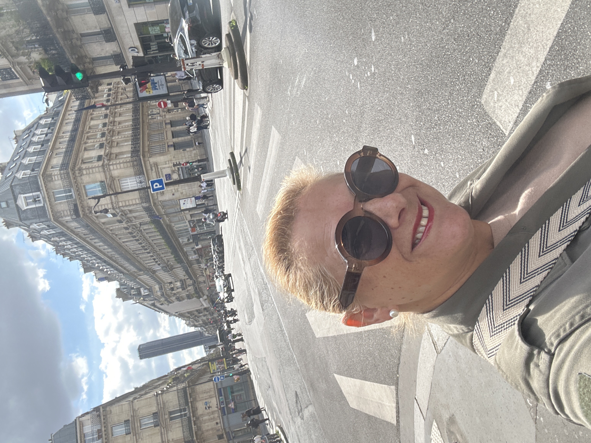 A medium shot shows a woman wearing sunglasses and a light-colored jacket with a patterned strap. The background features a city street with buildings, cars, pedestrians, and a crosswalk. The sky has clouds, and the building's architecture suggests a European city. A tall building is visible further down the street.