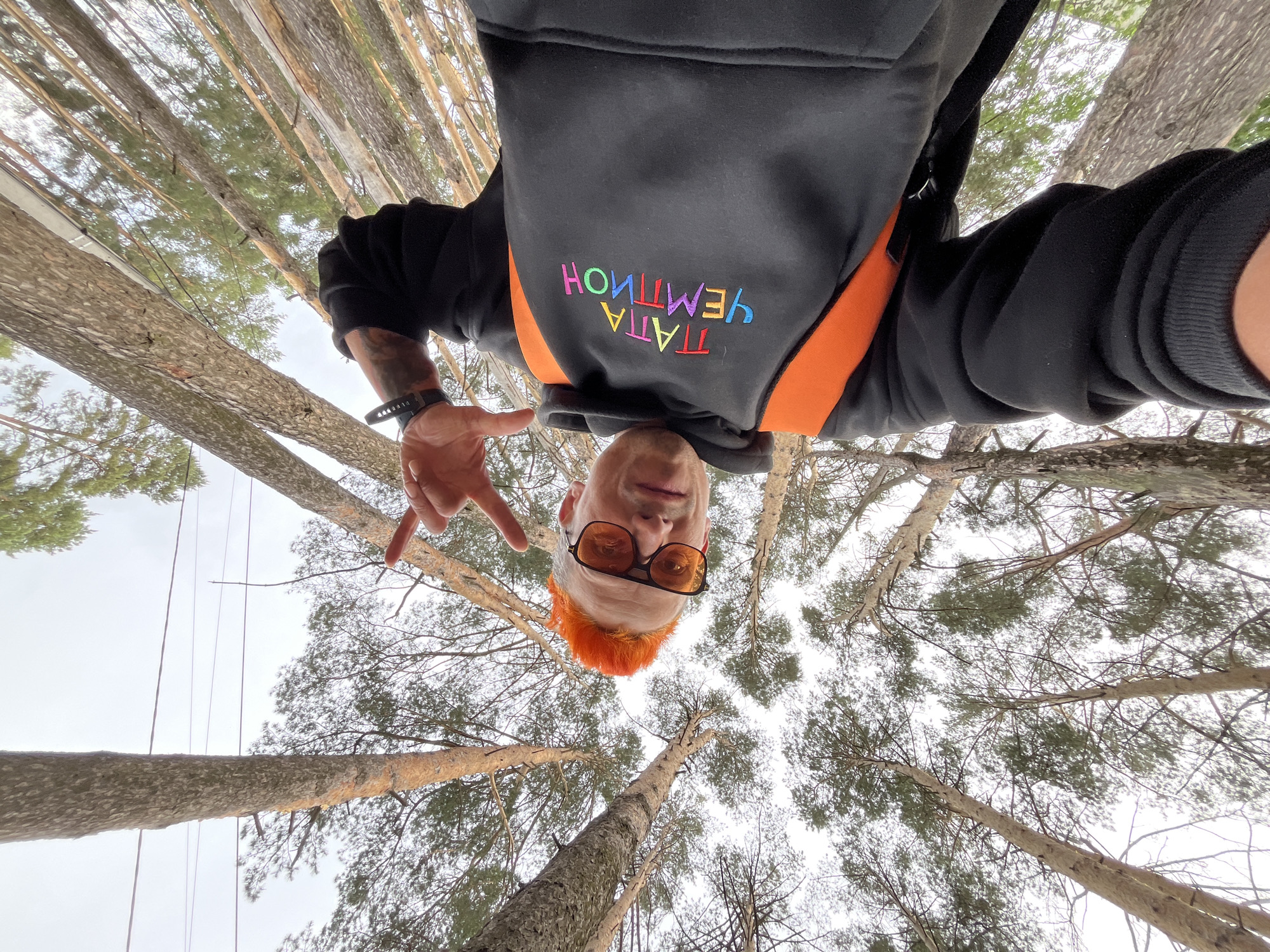 A man with orange hair and amber-tinted glasses is taking a selfie from a low angle, looking down at the camera. He wears a black hoodie with a colorful Russian phrase stitched onto the chest and an orange strap visible across his shoulder. His left hand is raised, with his index finger extended. The scene is set in a forest near Chaikovsky, Russia, suggested by the surrounding tall pine trees reaching towards the overcast sky. The perspective indicates the camera is positioned close to the ground, pointing upwards.