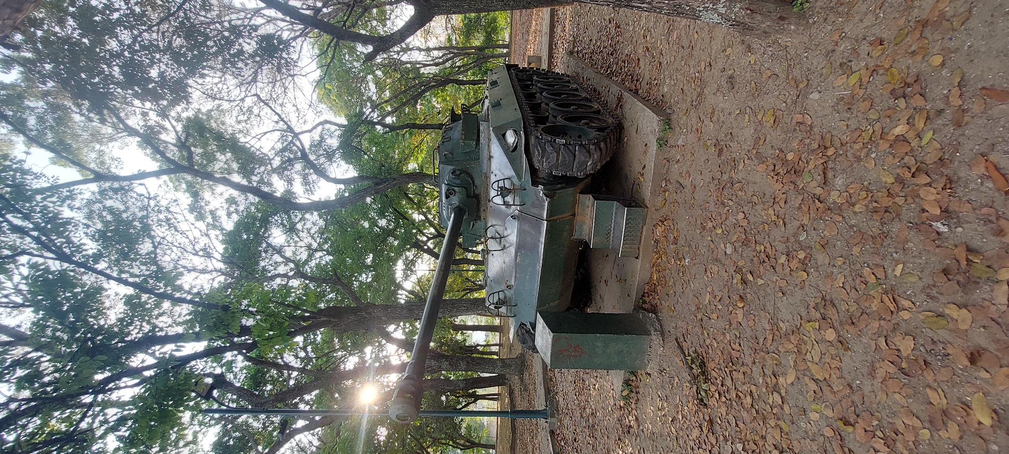 The image depicts a World War II-era tank on display at a park in Caracas, Venezuela. The tank, likely a Sherman or similar model, is painted in military green and sits on a concrete base. The surrounding environment is a wooded park area with trees and fallen leaves on the ground. The tank's gun is pointed towards the left side of the frame, and sunlight can be seen filtering through the trees in the background. There is no presence of people in the image.