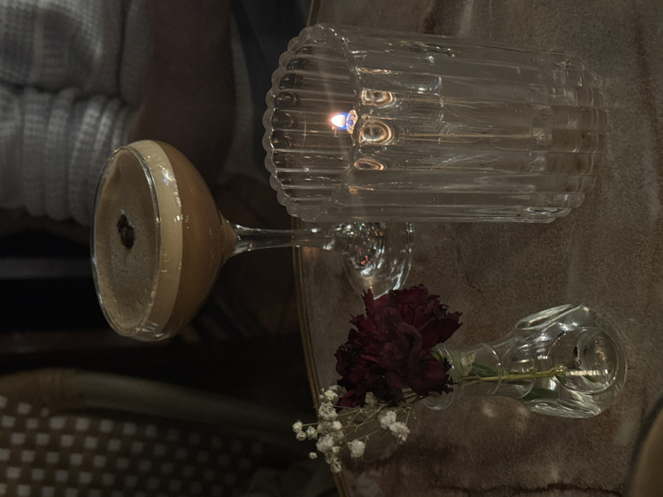 A martini glass with a coffee-colored drink rests alongside a ribbed glass candle holder with a lit candle. Nearby, a small glass vase holds a deep red carnation and sprigs of white baby's breath. All items sit on a textured surface. A blurry light-colored sweater is in the background.