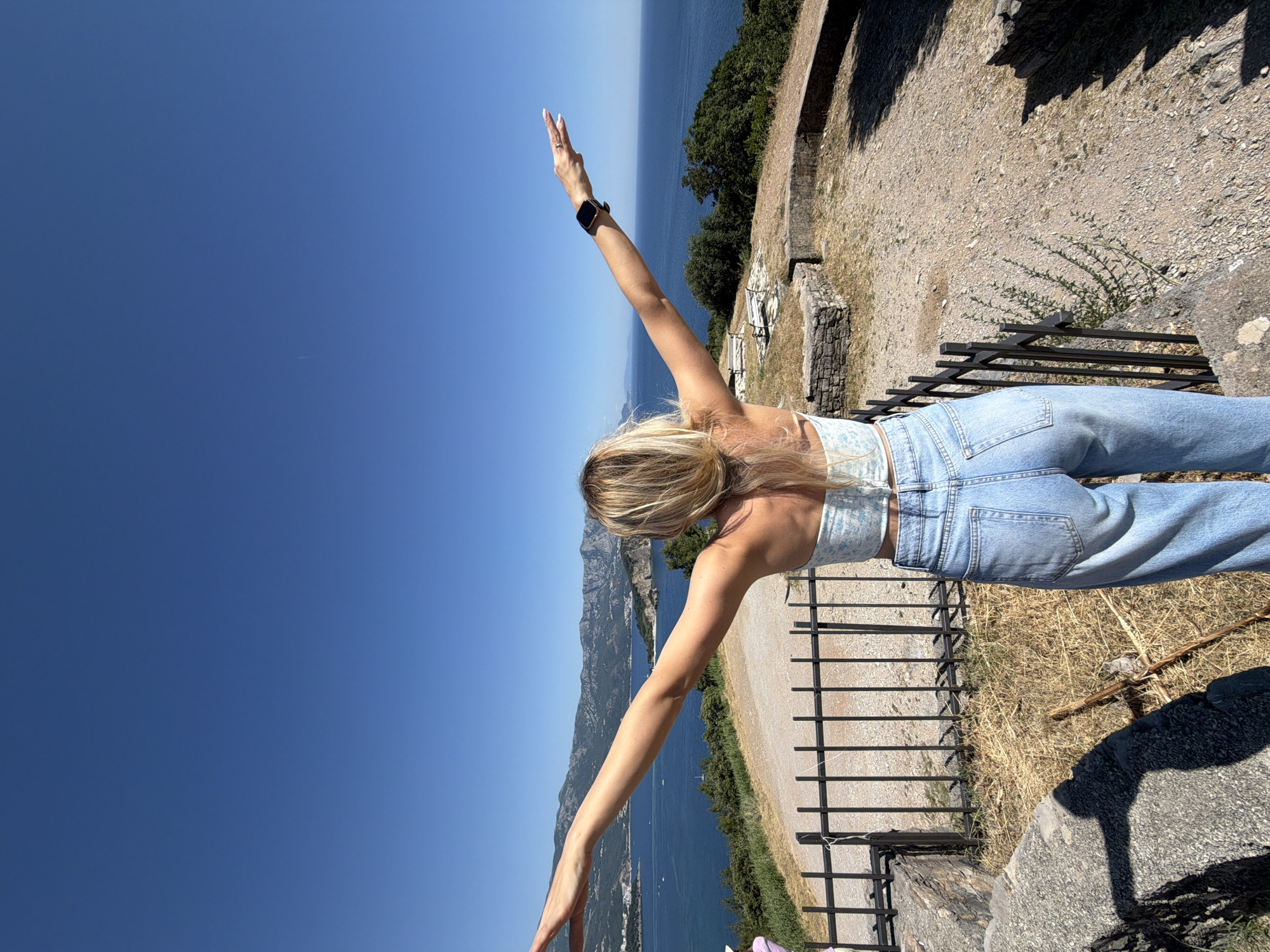 A female individual is depicted from a rear perspective, standing on an elevated, rocky, and dry dirt ground. She possesses long, blonde hair and wears a light-colored strapless crop top with a pattern, paired with light-wash blue jeans. Her arms are outstretched horizontally, with a dark-colored smartwatch visible on her left wrist and a ring on her left hand.
In the immediate foreground, a black metal fence separates the viewer's current position from the edge of the viewpoint. To the left, a large grey stone structure is partially visible. To the right, low stone walls and foundations, consistent with ancient ruins or an observation platform, are present on the dry ground. Sparse patches of dry grass and small green bushes are scattered across the foreground terrain.
Beyond the fence, a panoramic coastal landscape unfolds. A vast expanse of clear blue sea occupies the mid-ground. To the left, a distant coastal town, identifiable as part of Budva, Montenegro, stretches along the base of large, verdant mountains under a cloudless, bright blue sky. Several boats are visible on the water. A prominent peninsula or island juts into the bay from the left. To the right, the sea extends towards the horizon, bordered by tree-covered hills. Two light-colored benches are situated on a paved section of the elevated ground to the right of the woman. The sky above is uniformly clear and blue.
