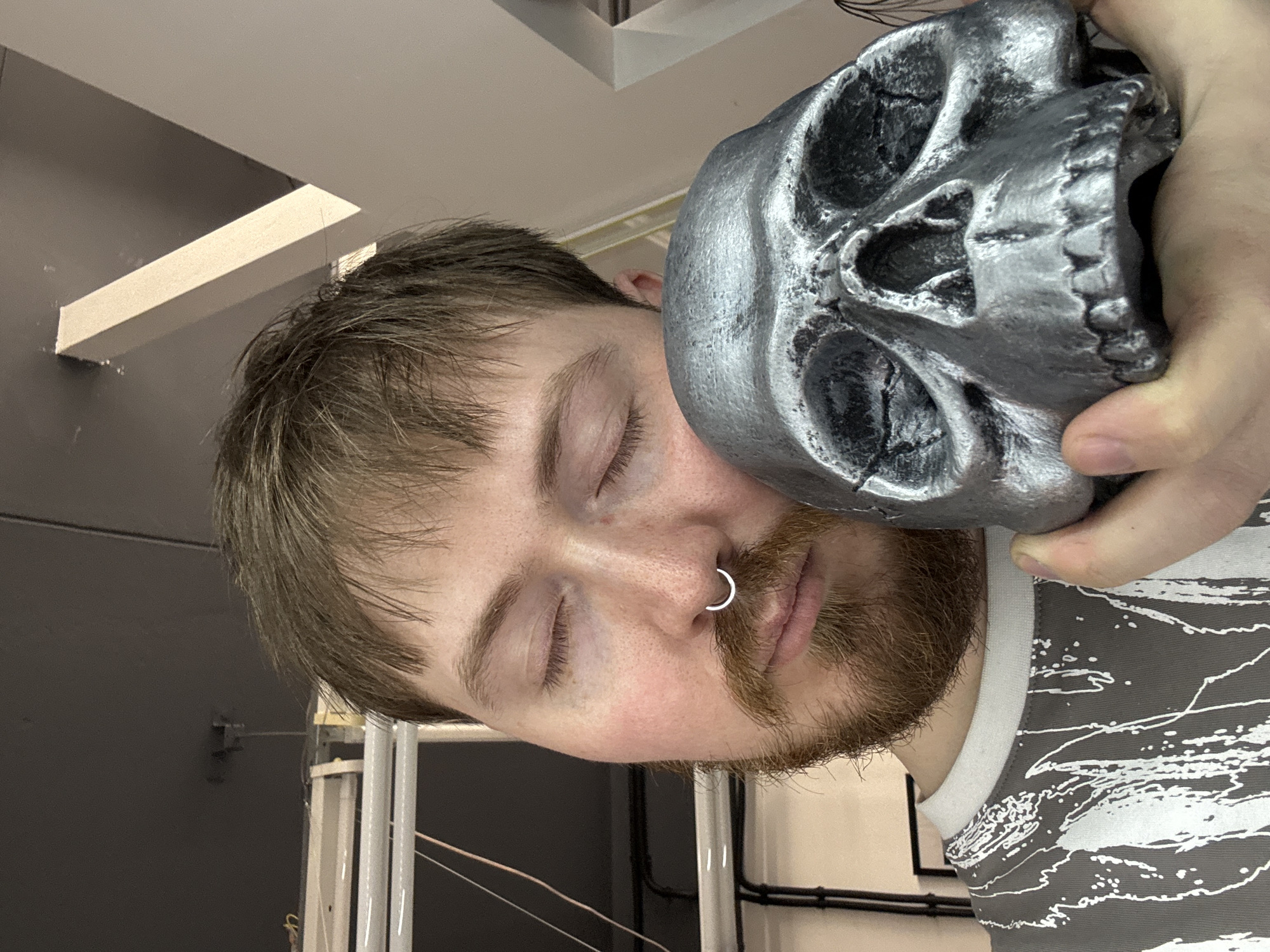 A medium shot depicts a man with closed eyes, holding a silver skull against his head. The man has light skin, brown hair with bangs, a beard, and a silver nose ring. He wears a gray and white patterned shirt. The silver skull is held in his right hand, obscuring part of his face. The background includes a gray wall and a white support structure, indicative of an interior space, with white beams near the ceiling. Based on the available information, the specific location within Chaikovsky, Russia, cannot be determined.