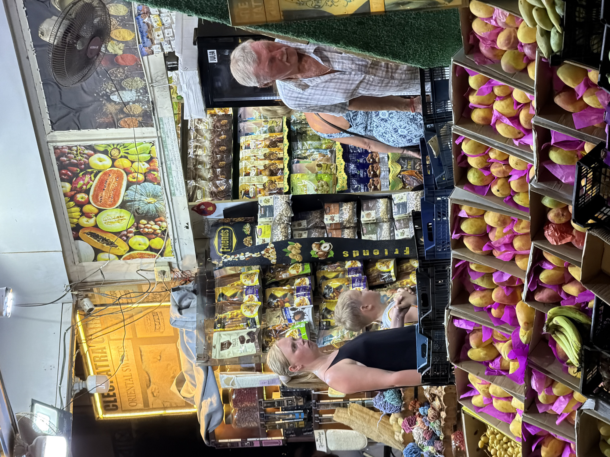 This image depicts a fruit and sweet stall in Sharm El Sheikh, Egypt. A Caucasian woman with blonde hair tied in a ponytail, wearing a black tank top, stands to the left of the frame. Beside her is a young Caucasian boy, likely her son, wearing a grey t-shirt. To the right, an elderly Caucasian man with gray hair, wearing a checkered short-sleeved shirt, is visible. The stall is filled with various packaged sweets and nuts on shelves. In the foreground, several crates are filled with ripe mangoes and other fruits, some wrapped in pink paper. Behind the stall, a colorful mural displays an assortment of fruits, and a ceiling fan is mounted above. A sign on the stall reads "Cleopatra Oriental Sweets." The lighting suggests it is nighttime or a dimly lit indoor market.