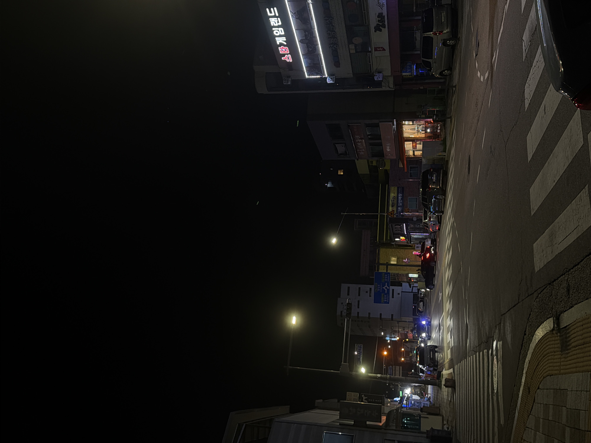 The image depicts a street scene at night in Dongducheon-si, South Korea. A street intersection is visible with multiple lanes of asphalt pavement marked with white lines. Pedestrian crosswalks are present, some with a raised tactile paving border. Streetlights illuminate the scene, casting a warm glow on the road and surrounding buildings.
On the right side of the street, a multi-story building displays a neon sign in Korean that reads "스타 게임랜드" (Star Game Land). Below this sign, there are windows and what appears to be storefronts. Several vehicles are present on the road, including a silver minivan, a dark-colored car, and a vehicle with its headlights on in the distance. Some cars are parked along the side of the street.
On the left side of the street, more buildings are visible, some with illuminated windows and signs. A lamppost with multiple lights stands near the sidewalk. A trash receptacle is also present on the sidewalk. The sky above is dark, indicating nighttime. The overall atmosphere is urban and active, with the lights of businesses and vehicles creating a vibrant nighttime environment.