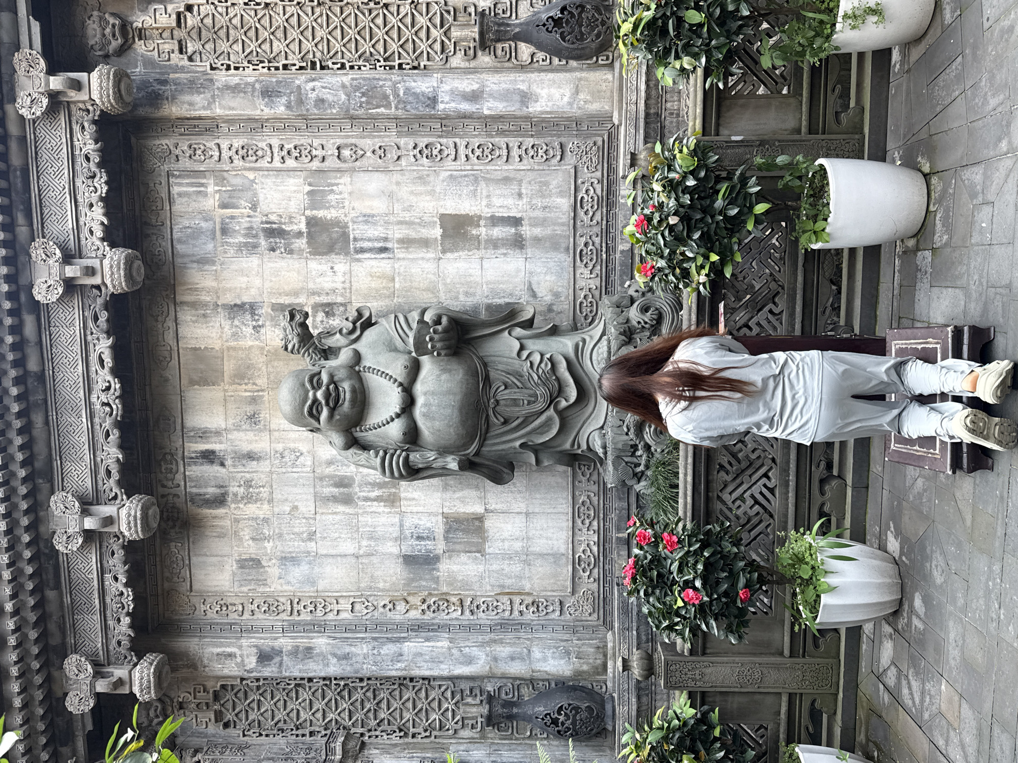 This image, rotated 90 degrees clockwise, presents an outdoor scene in what appears to be a traditional Chinese architectural setting, likely a temple or courtyard in Yuzhong District, China. The central subject is a large, gray stone statue of a smiling, corpulent figure, characteristic of Maitreya Buddha (Budai or Hotei). The statue is intricately carved, depicting the figure with a beaded necklace and flowing robes.
To the right of the statue, a person with long brown hair, seen from behind, stands observing the statue. They are dressed in casual, light gray attire, consisting of a long-sleeved top and pants, paired with light-colored sneakers.
The background features a highly detailed, dark gray stone wall or facade. It is adorned with elaborate carvings, including geometric patterns, lattice designs, stylized cloud or floral motifs, and decorative borders. Ornate architectural brackets or supports are visible along the upper and left edges of the wall. A dark, vase-like carving is also embedded in the wall on the right side.
Along the right side of the frame, several potted plants in large white planters add greenery and color to the scene. Some plants display lush green foliage, while others feature vibrant red flowers, possibly camellias. The ground is paved with dark gray stone tiles. The lighting suggests daytime, with soft, indirect light. There is no visible text in the image.