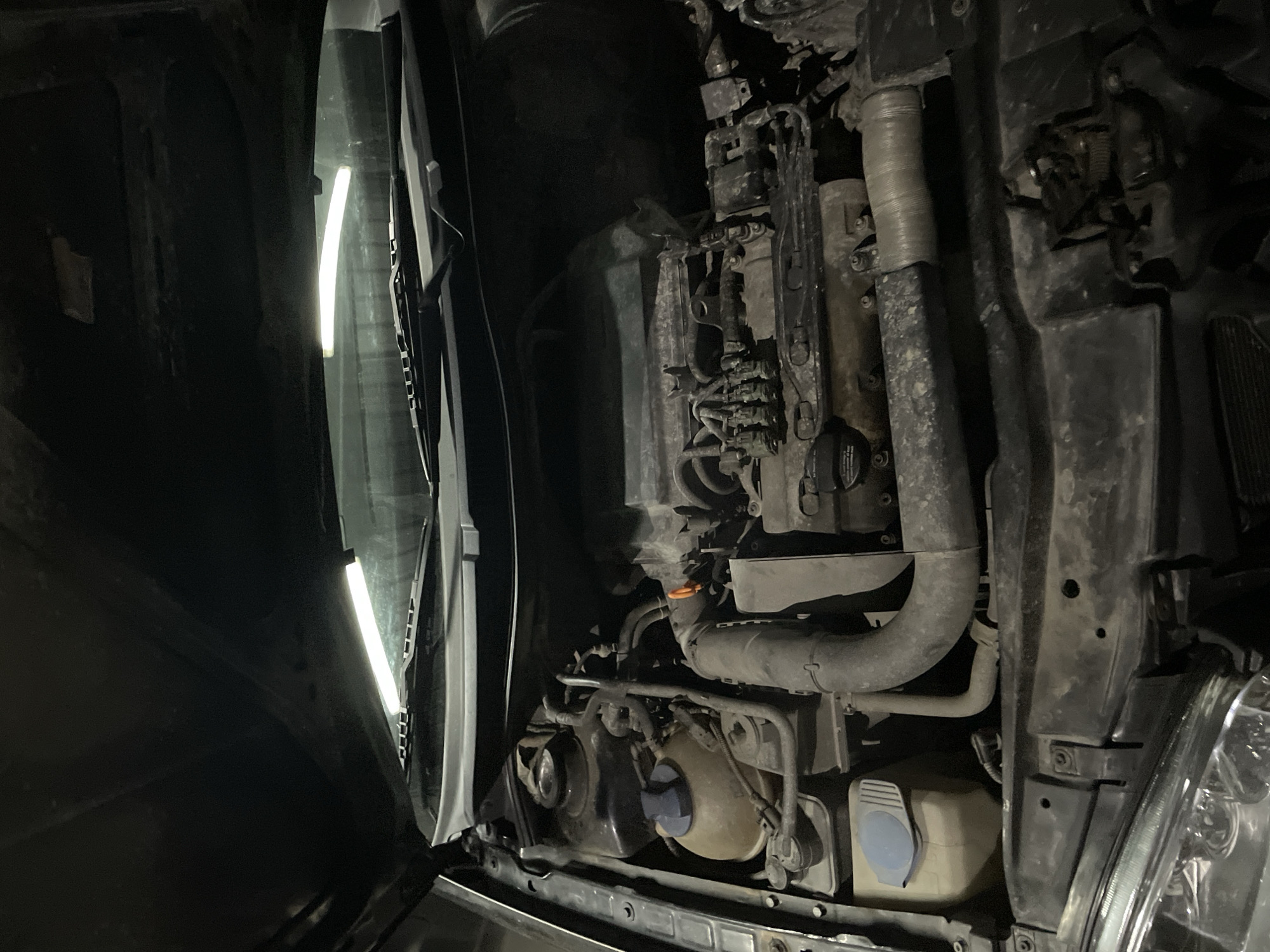 The image shows the engine compartment of a vehicle. The hood is open. The engine components are visible, including hoses, fluid reservoirs with blue caps, and engine parts covered in grime and dirt. A work light illuminates the left side of the engine bay. The location is likely outdoors, based on the dim ambient light. The presence of windshield wipers and headlight components suggests the front of the vehicle is facing the viewer. There is no indication of the specific location within Zlatograd, Bulgaria, nor is there any evidence of people or actions taking place.