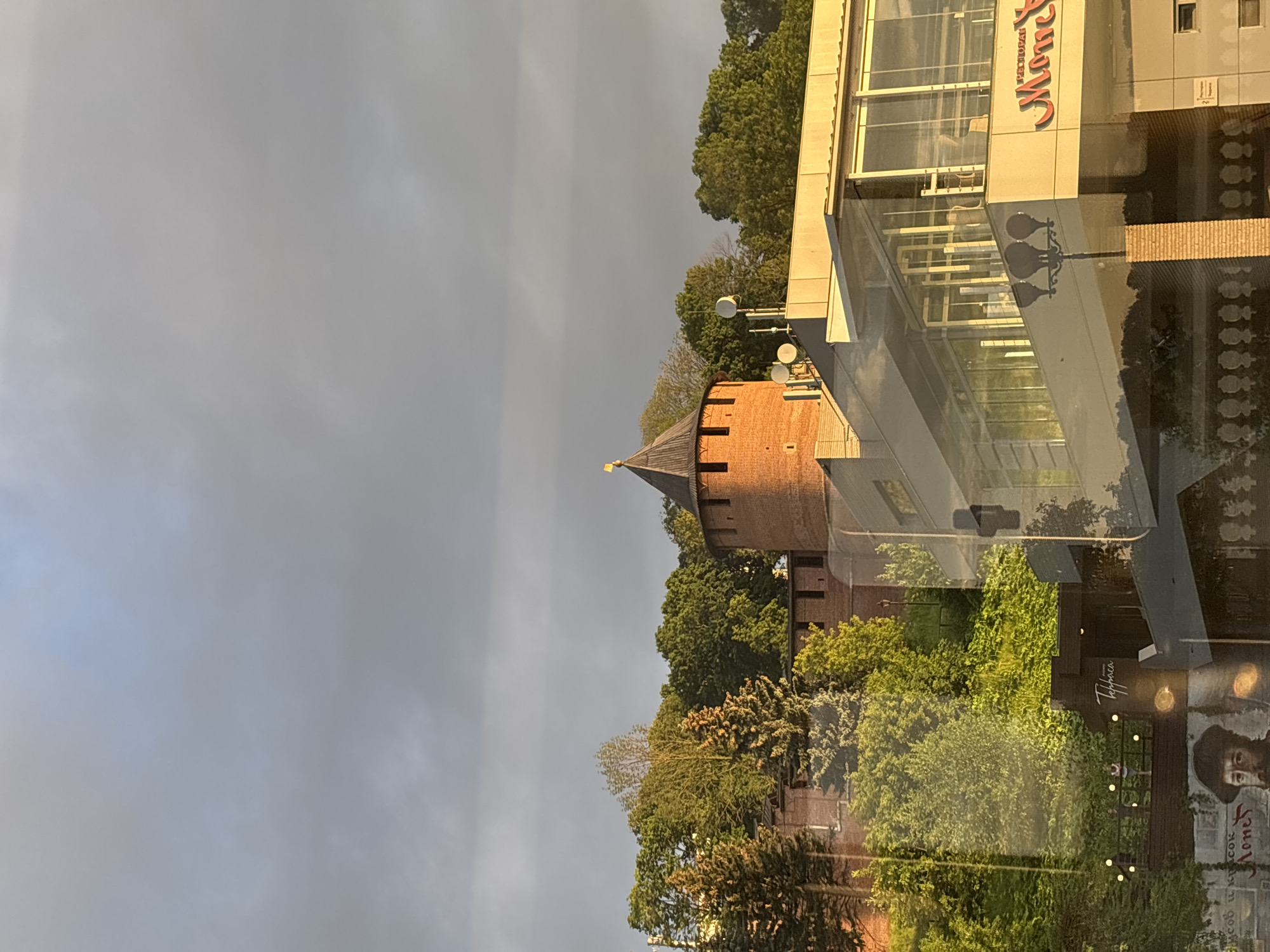 The image shows a view of the Nizhny Novgorod Kremlin, a historic fortress in Nizhny Novgorod, Russia. A round brick tower with a conical roof and a golden finial is visible against a cloudy sky. In the foreground, a modern building with large glass windows and a sign that reads "Moments" is partially obscuring the view of the Kremlin walls and lush green trees. The lighting suggests it is late afternoon or early evening, with the sun casting a warm glow on the modern building. A lamppost is also visible in front of the modern structure.