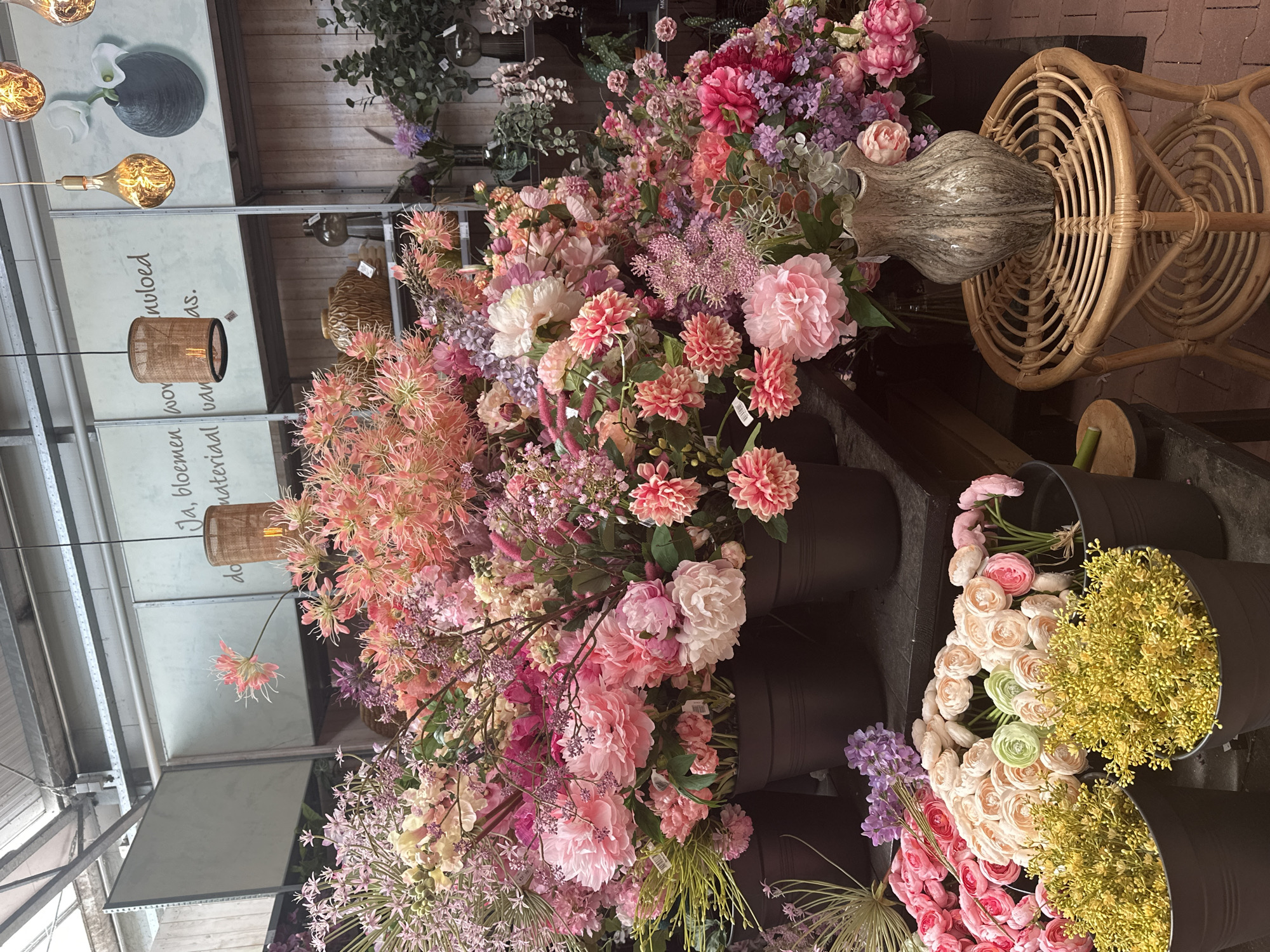 This is an eye-level, vertical, medium shot of a vibrant display of artificial flowers in what appears to be a store or market in Tuk, Netherlands. The subjects are an abundance of various artificial flowers in shades of pink, purple, yellow, and white. The flowers are arranged in large groupings, some in dark buckets or containers, creating a lush and abundant visual.
Notable objects include several large, dark, cylindrical buckets holding the flowers, a woven rattan chair with a circular design, and a tall, decorative vase with a marbled gray and white pattern placed on the chair. Hanging from the ceiling, illuminated by light bulbs, are at least two pendant lamps with woven shades, and another large, Edison-style bulb hangs by a wire.
The setting appears to be indoors, possibly a greenhouse or flower shop, with a wooden plank ceiling and exposed metal beams of a larger structure above. There are also indications of shelving or displays in the background.
Visible text includes "Ja, bloemen voer" and "materiaal van" on a light-colored panel in the background, which seems to be promotional material for the flowers. There are also small white tags on some of the flower stems, typical of retail items.
The overall impression is one of a colorful and abundant floral display, likely aimed at attracting customers. The lighting suggests it could be daytime, though the exact time of day is not discernible. There are no people or animals visible, and the focus is entirely on the floral arrangement and associated decorative objects.