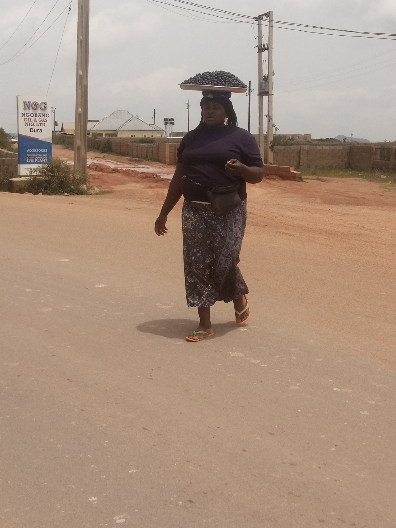 In Jos, Nigeria, a woman walks on a paved road carrying a tray of dark, round objects on her head. She is wearing a dark purple t-shirt, a patterned skirt, and flip-flops. A black fanny pack is worn around her waist. She appears to be holding something small in her right hand. The background shows a sign for "NOG NGOBANG OIL & GAS NIG. LTD. Dura" and a few buildings. The sky is overcast.