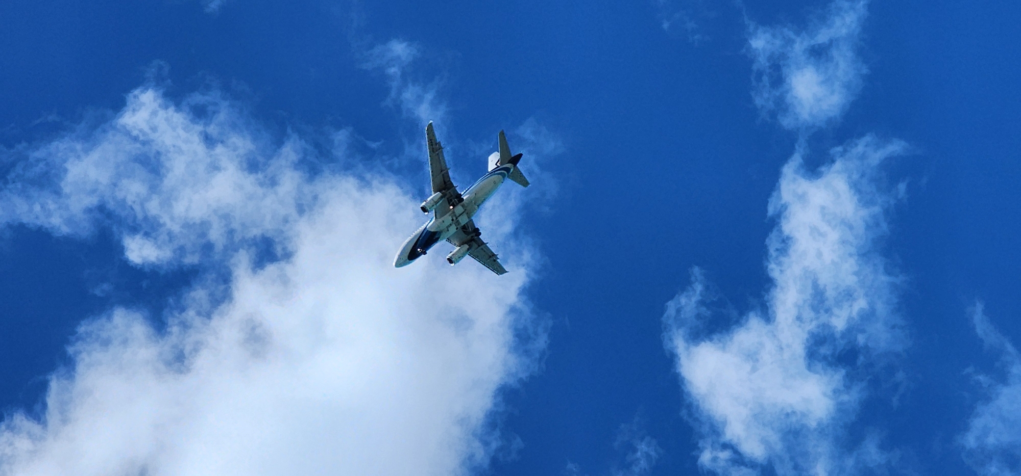 A commercial airplane is captured from below, ascending into a bright blue sky filled with scattered white clouds. The aircraft is white with blue accents and appears to be in active flight, with its landing gear retracted. The scene is set against a clear blue sky, characteristic of tropical weather. The image is taken from ground level looking upwards, emphasizing the vastness of the sky and the scale of the airplane. No people are visible. The location is specified as Baan Chaweng Noi, Thailand.