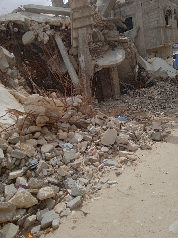 The image displays a scene of significant structural destruction in Khan Yunis, Palestinian Territory. A multi-story building has suffered extensive collapse, with its upper sections reduced to rubble and a substantial portion of the facade missing. Exposed, twisted rebar protrudes from concrete slabs and fragmented columns, indicating the severity of the damage. A large pile of mixed debris, consisting of broken concrete blocks, shattered masonry, and dust, extends across the foreground and midground. Remnants of a partially standing wall, constructed from grey concrete blocks, are visible on the right side, featuring dark, empty window openings. In the lower midground, amidst the rubble, a crumpled red and white can and a blue plastic object, possibly a tarp or bag, are present. The immediate foreground consists of a sandy dirt path. The sky is overcast. No individuals or ongoing activities are visible.