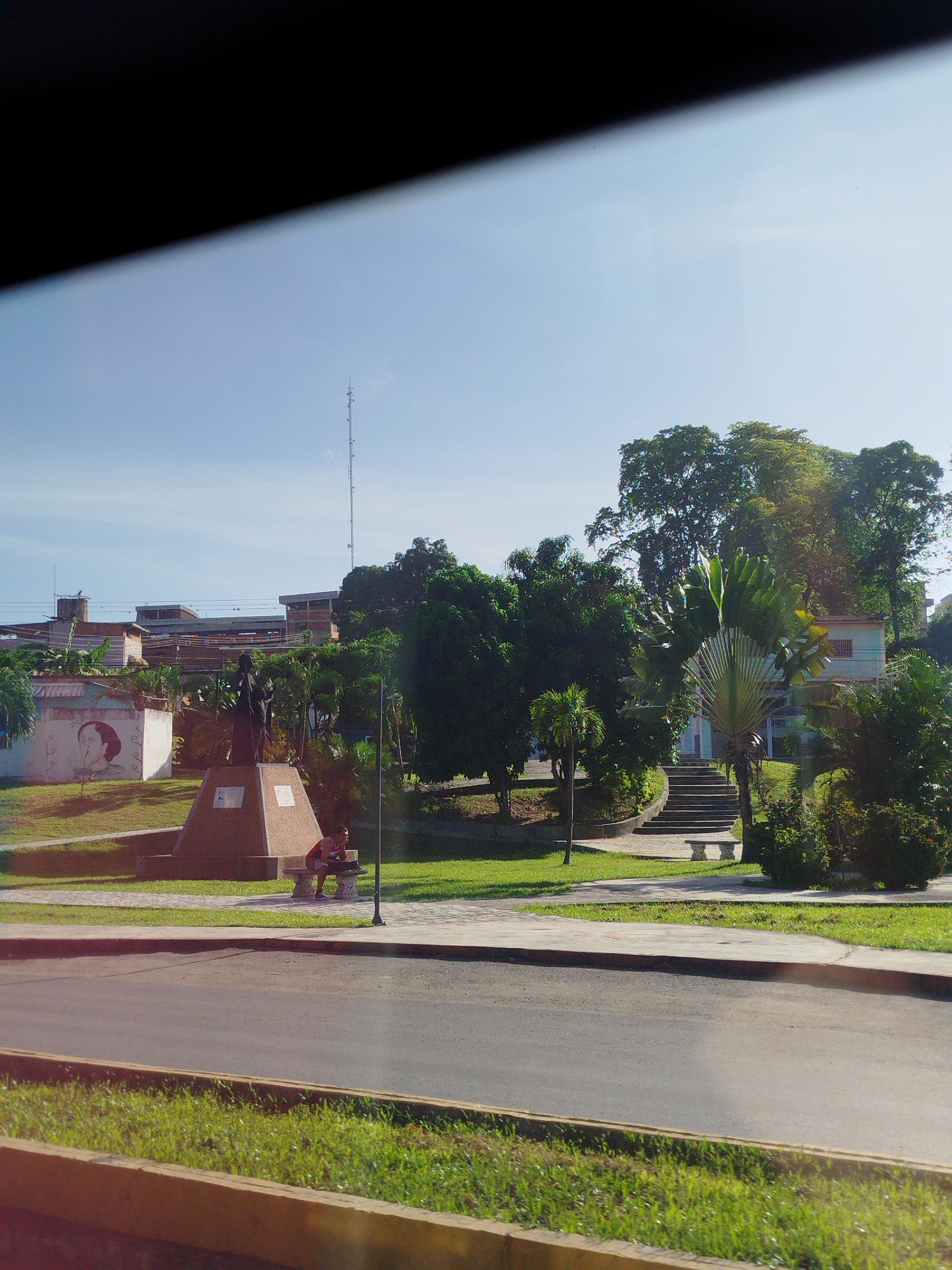 The image depicts a sunlit outdoor scene in Parroquia Charallave, Venezuela, featuring a monument in the foreground, lush greenery, and buildings in the background. A dark, imposing monument with a pyramidal base stands prominently in the center, topped by a statue of two figures. To the right of the monument, a person is seated on a bench, looking away from the camera.
Behind the monument, a white building is visible with a large, circular mural of a woman's face on its exterior wall. Further back, a complex of multi-story buildings with visible scaffolding and exposed brickwork suggests ongoing construction or renovation. A tall communication tower rises above the structures in the distance.
The environment is characterized by well-maintained grassy areas and a paved pathway leading up a set of stairs. Several types of trees and tropical plants, including a large fan palm, contribute to the verdant atmosphere. A paved road borders the park area, with a strip of grass and a yellow concrete border separating it from the park. The sky is clear and bright blue, indicating fair weather. The overall impression is one of a public space within a town or city, likely a park or plaza, given the monument and seating.