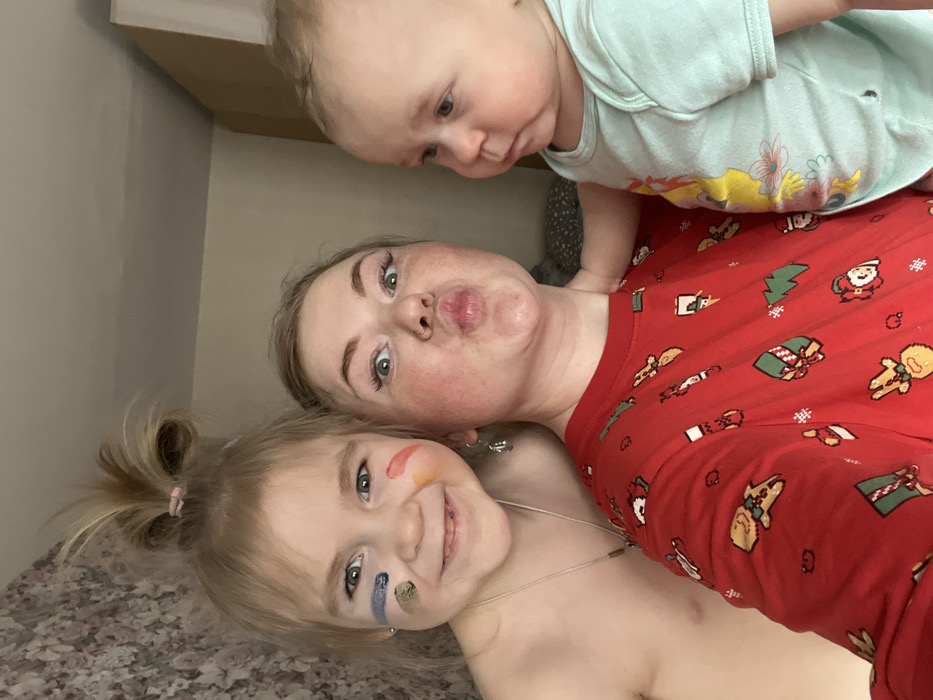 An indoor selfie features three light-skinned individuals: an adult female, a young female child, and an infant. The adult female, positioned centrally, has light brown hair, dark eye makeup, a nose piercing on her right nostril, and a silver-colored earring in her left ear. She wears a red long-sleeved shirt with repeating patterns of gingerbread men, Christmas trees, presents, reindeer, snowmen, and stockings, and is looking at the camera with wide eyes and pursed lips. To her left, a young female child, approximately 3-5 years old, has blonde hair tied in a top knot with a pink elastic. She smiles directly at the camera, displaying blue and green face paint stripes near her left eye and red and orange stripes near her right eye. She wears a thin silver-colored necklace and is bare-chested. To the adult's right, an infant, appearing less than one year old, wears a light blue short-sleeved bodysuit with a partial yellow graphic and looks towards the right side of the frame with a neutral expression. The adult female's right arm is partially visible around the infant. The background includes a light-colored wall with a subtle floral wallpaper pattern on the left and a plain light-colored wall or ceiling segment on the right, along with a white vertical object, indicating a domestic interior. The specific location within Chaikovsky, Russia, cannot be determined from the image.