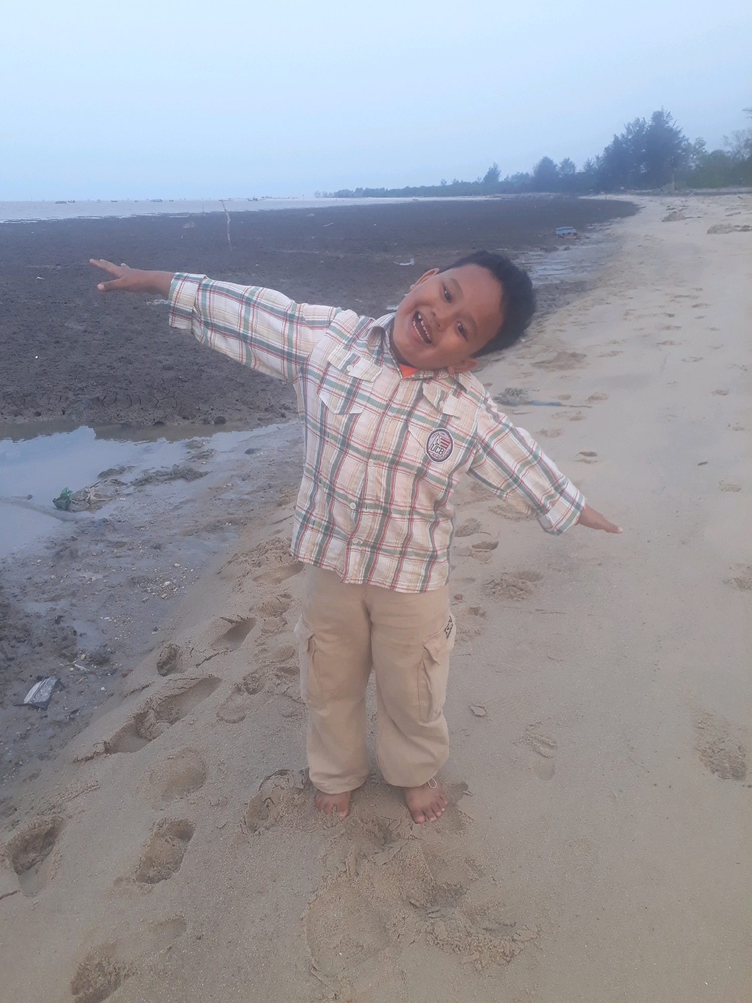 A young boy stands on a sandy beach, his arms outstretched to the sides with his body leaning back and head tilted. He is wearing a plaid shirt and khaki cargo pants. The beach appears to be a low tide area with mudflats visible in the background. The sky is overcast and the overall lighting is dim. There are footprints in the sand and some debris on the mudflats. The location is identified as Unknown, Indonesia.