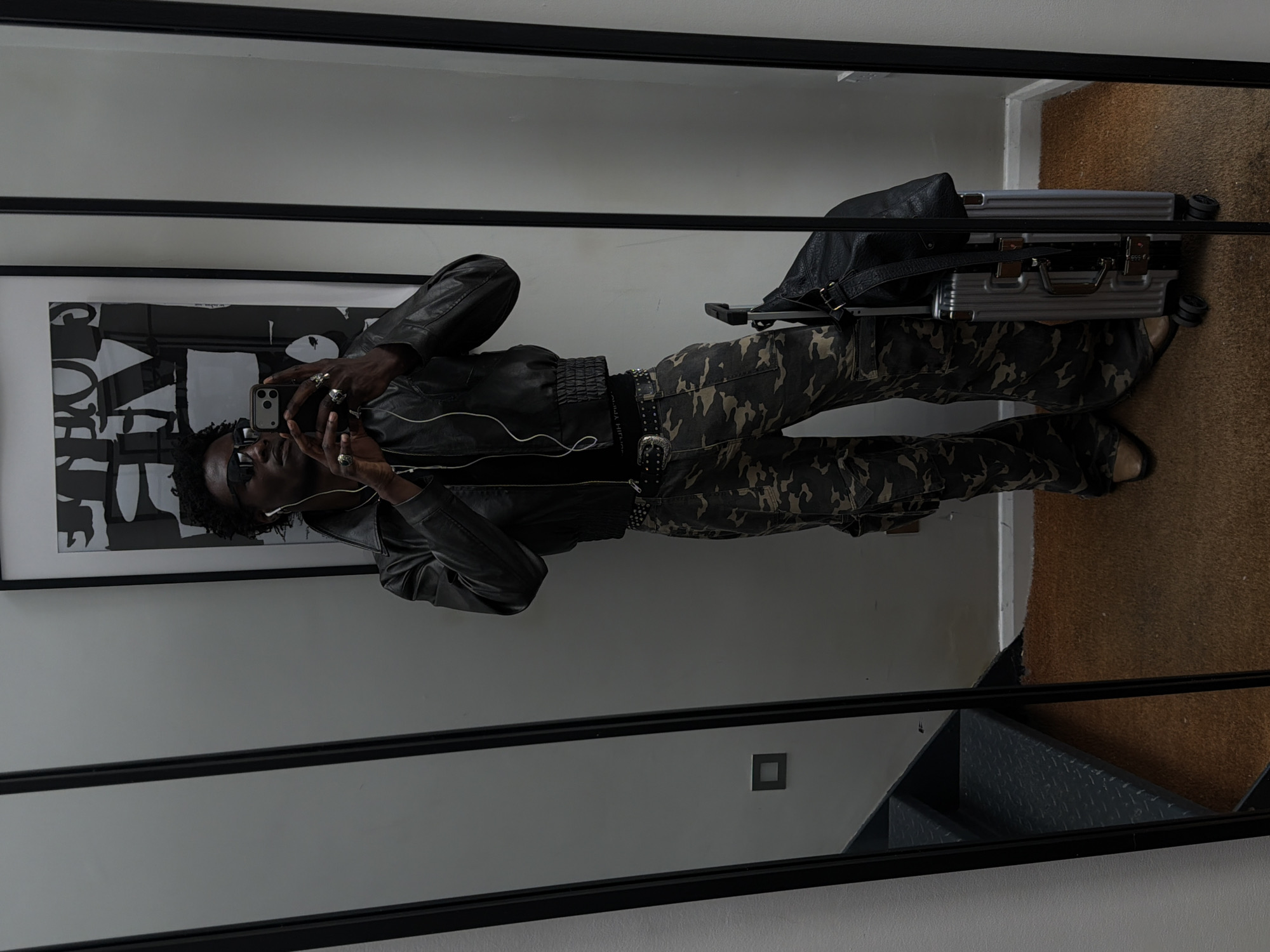 A person is taking a selfie in a mirror. The person is wearing a black leather jacket, camouflage pants, a studded belt, and sunglasses. They are also wearing headphones. A framed black and white print hangs on the wall next to the mirror. A silver suitcase and black leather bag are on the floor. The mirror is framed in black. Below the mirror, a staircase with diamond-patterned metal treads is visible. The location appears to be an apartment or home interior in London, United Kingdom.