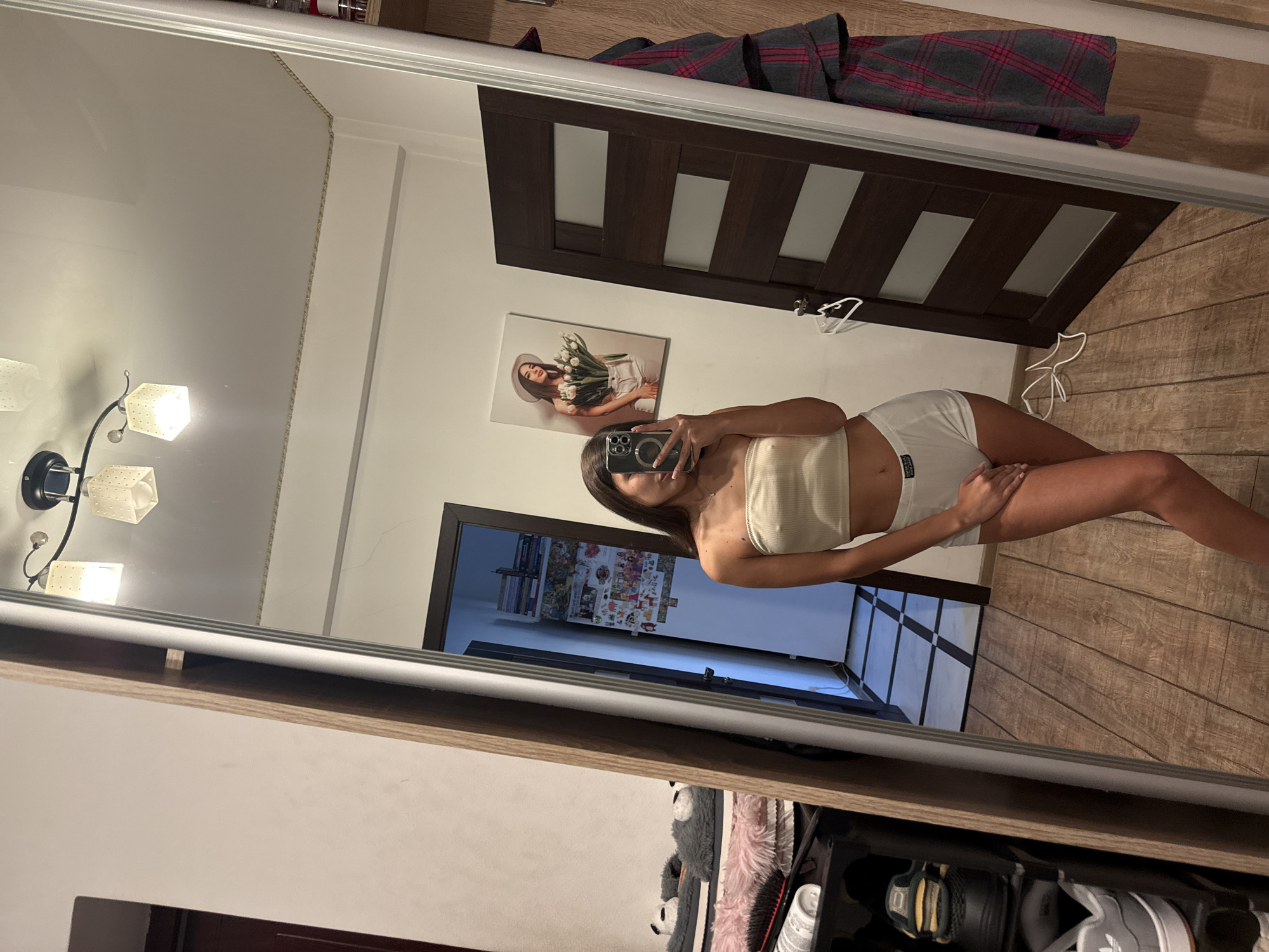 An interior full-length mirror selfie shows a young woman with long brown hair, wearing a cream-colored strapless bandeau top and white boxer briefs. She is standing in what appears to be a bedroom. The mirror reflects the room, revealing a door with horizontal wood panels, a framed picture of a woman holding tulips on the wall, and a shoe rack filled with shoes. A floor-length mirror is positioned to the left of the frame, reflecting a bed with fuzzy grey slippers and pink fluffy pillows. The lighting suggests it is daytime. The scene appears to be a private residence, likely a bedroom, and there is no indication of the location being in Chișinău, Moldova.