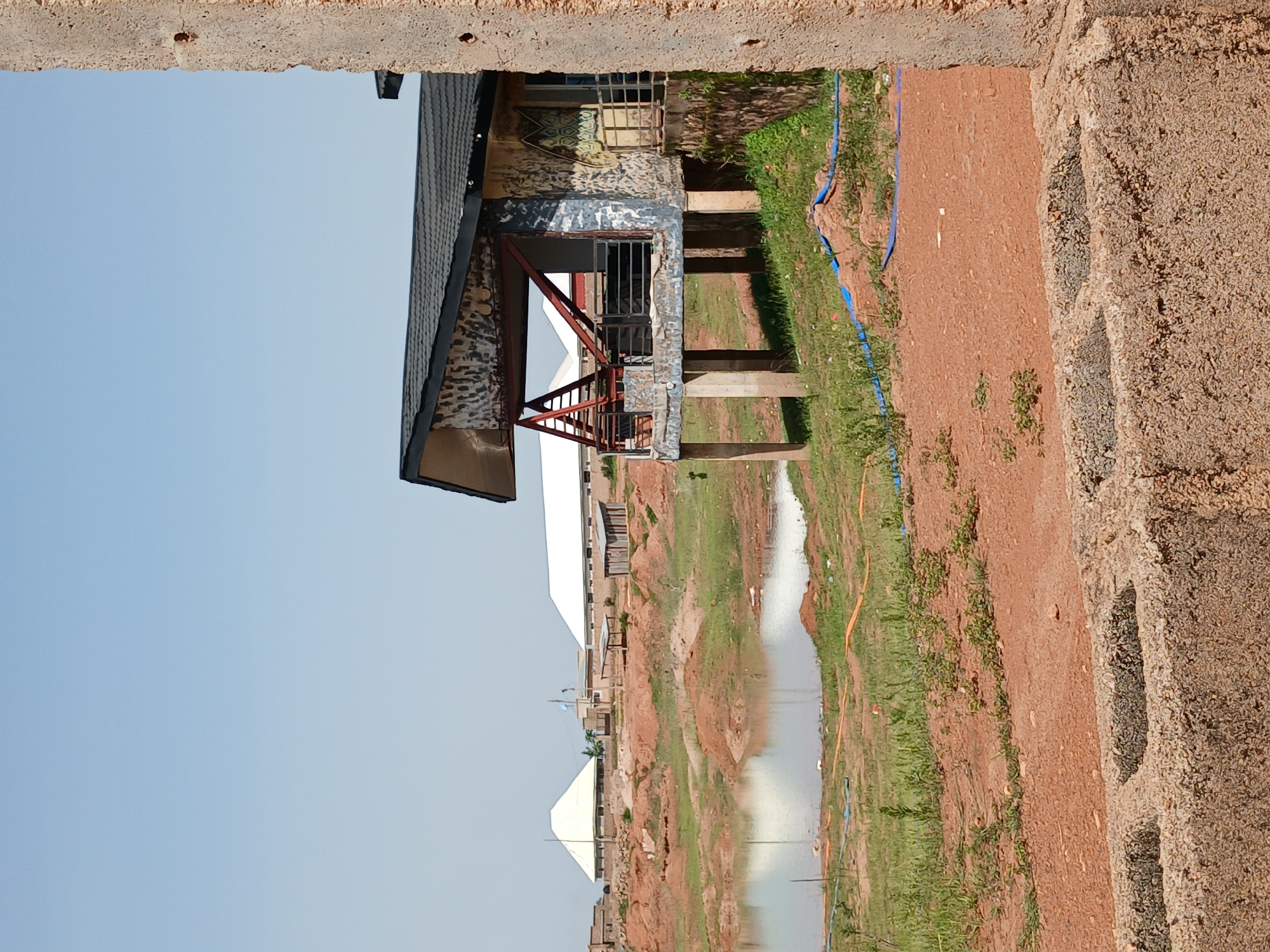 The image depicts an outdoor scene, likely in Kwang, Nigeria, featuring a partially constructed or weathered building with a dark, pitched roof supported by concrete pillars. The building has a balcony with metal railings and a patterned facade. In the foreground, a wall made of concrete blocks is visible. Behind the building, there is a body of water, surrounded by disturbed earth and vegetation. Further in the background, several white-roofed structures, possibly buildings or tents, are present. The sky is clear and pale blue. The overall impression is one of an unfinished or developing construction site.