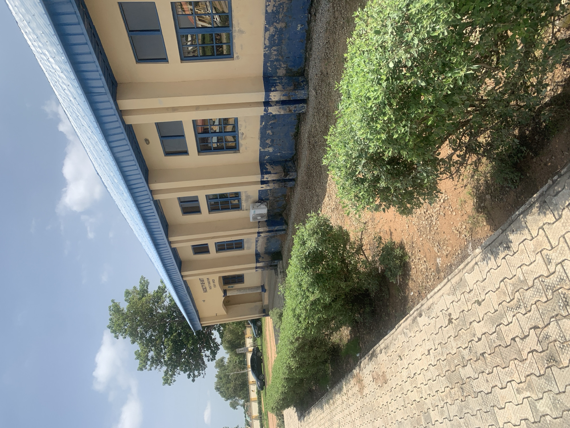 The image depicts the Faculty of Art building at a Nigerian university, possibly Taraba State University in Jalingo, Nigeria. The building has a beige facade with a blue, corrugated metal roof and blue-framed windows. The lower portion of the building exhibits wear and tear, with chipped blue paint revealing a lighter colored base. In front of the building, a paved walkway runs alongside a row of neatly trimmed green bushes. Beyond the bushes, there is a dirt area with some gravel. Two dark-colored cars are parked in the background, and trees and other vegetation are visible in the distance under a partly cloudy sky. A sign on the building indicates "FACULTY OF ART", "FACULTY BOARD ROOM", and "STAFF OFFICES". An air conditioning unit is mounted on the exterior wall of the building. The scene suggests a warm, sunny day.