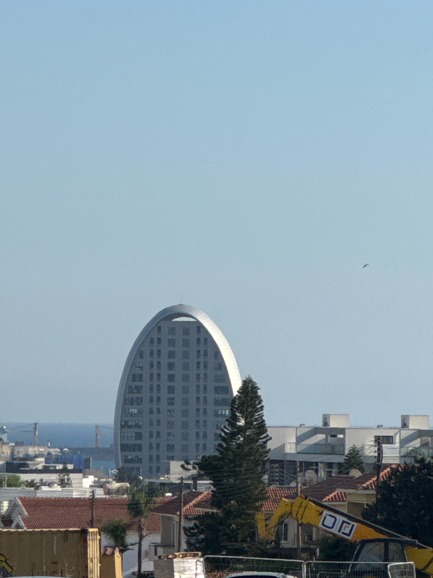 The image shows a cityscape with a distinctive arched building as the central focus. The building is tall and grey, with a grid-like pattern of windows. It is situated behind several lower buildings with red-tiled roofs. In the foreground, a yellow construction vehicle with the JCB logo is visible. A tall, dark green conifer tree stands to the left of the construction vehicle. The sky is clear and blue with a small bird flying in the distance. The scene appears to be in Agios Athanasios Municipality, Cyprus, given the architectural style of the buildings and the clear, sunny weather typical of the region. The overall impression is one of urban development with ongoing construction.