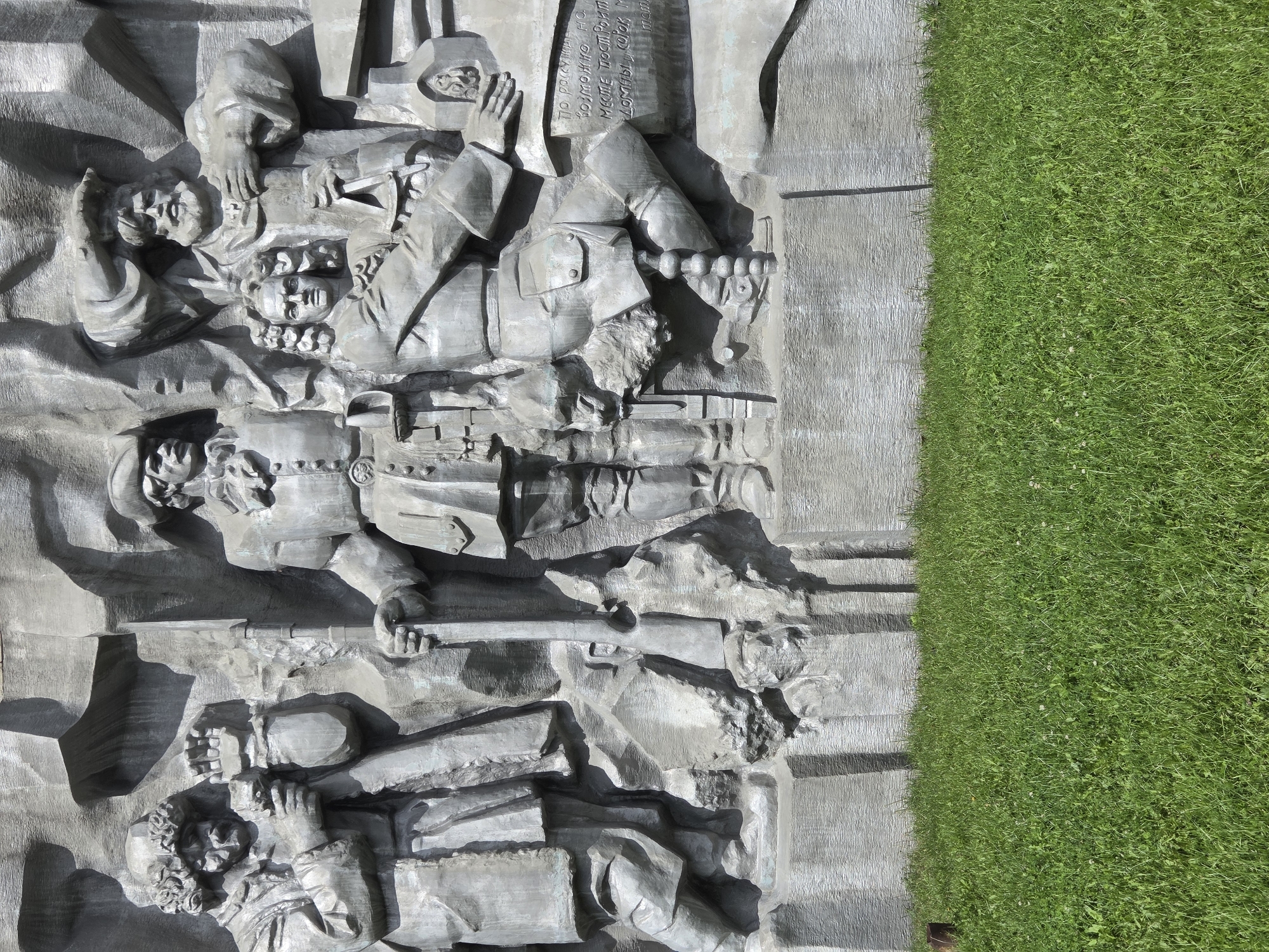 This is a detailed, low-angle, full shot of a bas-relief sculpture located in Yekaterinburg, Russia. The sculpture depicts a group of figures, predominantly male, in historical attire. The figures are carved in stone and are rendered in high relief, creating a sense of depth and volume.
On the far left, a figure with curly hair and a furrowed brow holds an object in his hand, possibly a tool or a document. He wears a long coat and appears to be in deep contemplation.
To his right, a central figure stands tall, clad in a military-style uniform with a hat. He holds a staff or a flag pole, his gaze directed forward.
To the right of the central figure, a younger man with curly hair is seated, holding a compass or a similar scientific instrument. He is dressed in formal attire, suggesting an intellectual or scholarly role.
Behind and to the right of the seated figure, a man with a long beard and a hat is visible. He appears to be gesturing with his hands, possibly in conversation or imparting knowledge.
The background of the sculpture is abstract and textured, suggesting rocks or rugged terrain. Inscriptions in Russian are visible on the lower right portion of the sculpture, though the text is partially obscured by shadow and weathering.
The sculpture is set against a backdrop of a rough, textured stone wall, and in the foreground, there is a strip of vibrant green grass. The lighting suggests it is a sunny day, casting distinct shadows across the relief, highlighting the textures and forms of the carving. The overall impression is one of historical significance, perhaps commemorating an event or individuals of importance.