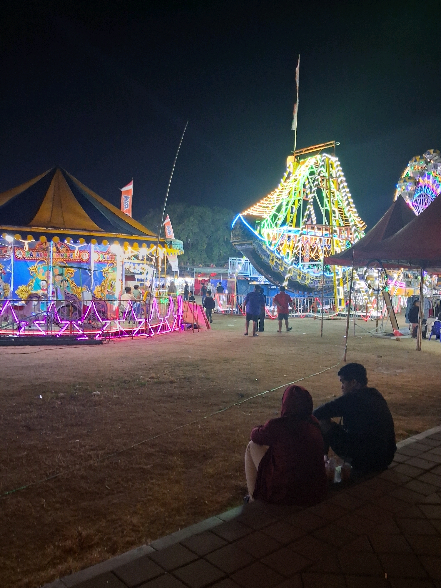At night, in a fairground area of Surakarta, Indonesia, a festive atmosphere is evident with illuminated rides. In the foreground, two individuals sit on the ground, facing away from the camera, towards the attractions. One person is wearing a dark hoodie and the other a maroon hoodie, with light-colored pants. The ground is covered with dry grass and a paved walkway is visible in the lower portion of the image.
Dominating the mid-ground are carnival rides. To the left, a colorful carousel with a yellow and black striped tent is lit with vibrant pink and blue lights. To the right, a large, elaborately decorated ride, possibly a Ferris wheel or a similar structure, is adorned with numerous strings of white, yellow, and green lights, outlining its intricate design. Several people are scattered around the fairground, some walking and others observing the rides. Flags are visible on poles, adding to the festive ambiance. In the background, trees are silhouetted against the dark night sky. Tents, likely for vendors or other attractions, are also present.