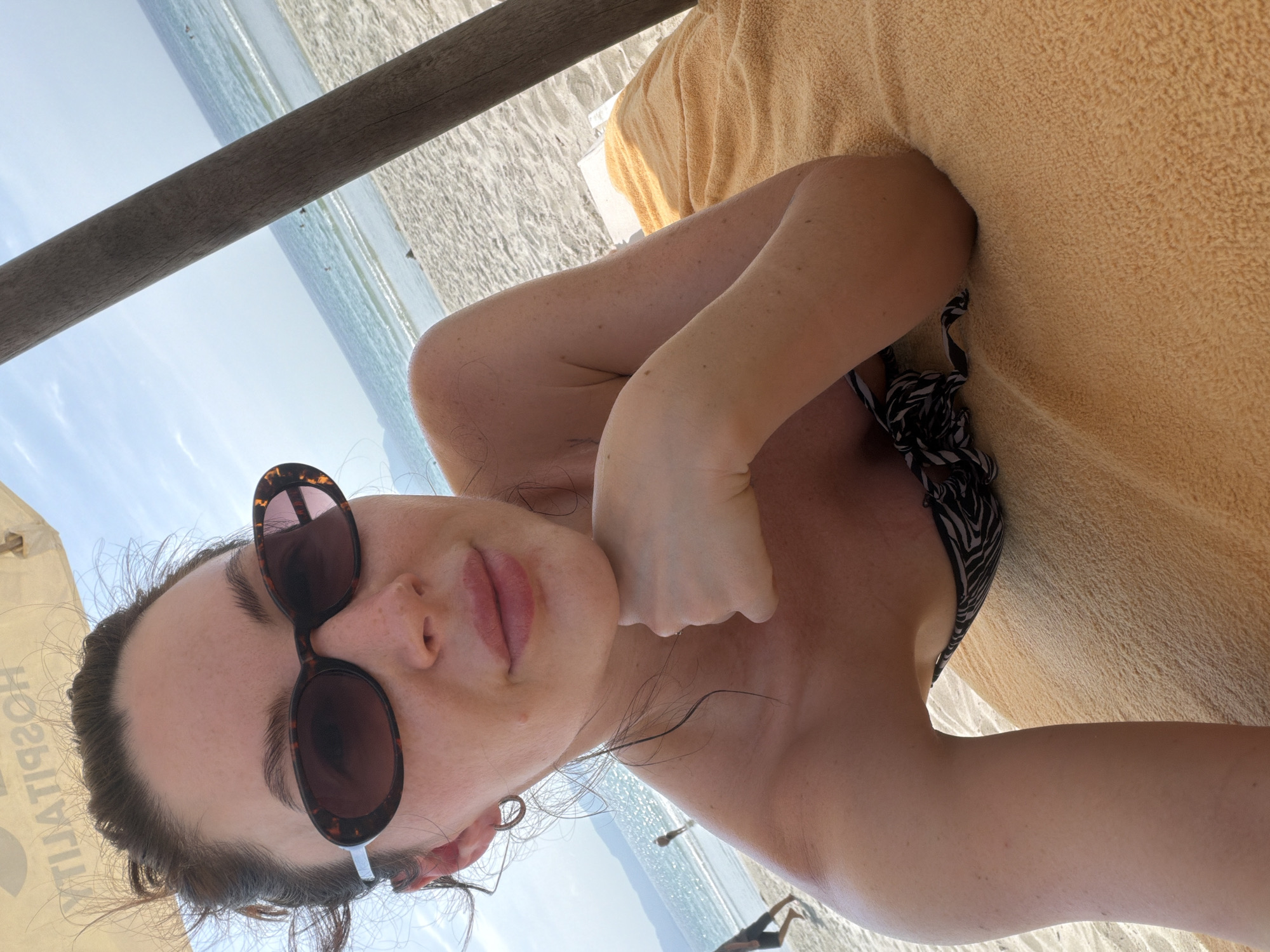 A woman lies on a beach towel on a sandy beach, resting her chin on her fist. She is wearing sunglasses and a black and white patterned bikini top. The ocean is visible behind her, with waves breaking on the shore and a few people in the water. A wooden post and a beach umbrella are to her right. The sky is clear and blue with a few clouds. The image is a selfie taken by the woman. The location is not identifiable as Dong Ninh Hoa Ward, Vietnam.