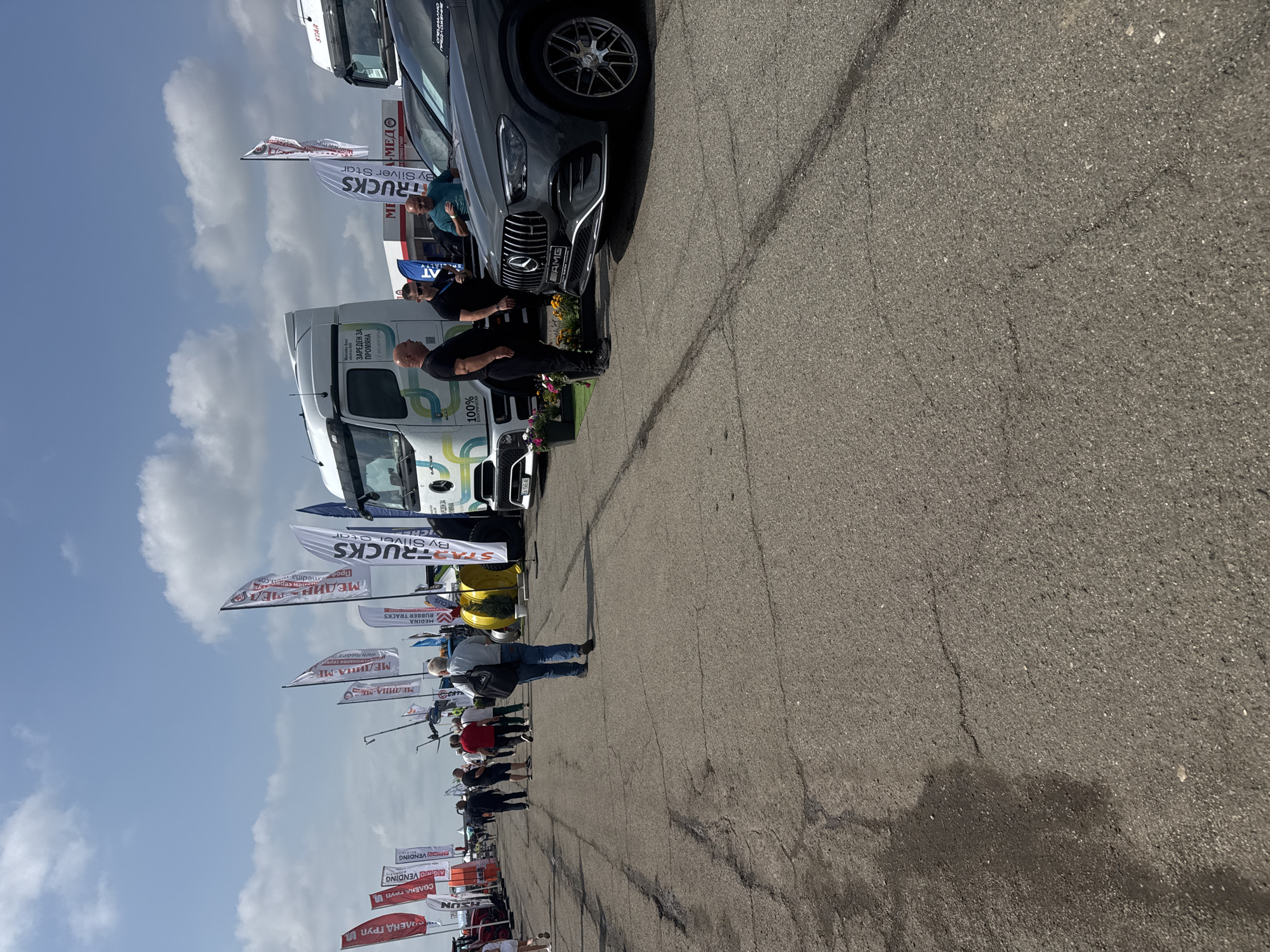 This media file captures an outdoor event or exhibition held on a wide, paved asphalt area, likely in Stara Zagora, Bulgaria. The scene is set under a partly cloudy sky during daylight hours.
The foreground features a dark gray Mercedes-AMG SUV prominently displayed, with two men standing near its front. One man, dressed in black, has his back to the viewer, while another in a black t-shirt stands closer to the vehicle. To the left, a large white Mercedes-Benz truck with green and black graphics, including "100%" and "ЗАРЕДЕН ЗА ПРОМЯНА" (Charged for change), is parked. Small decorative flowers are placed near the front of the SUV.
The event appears to be a trade show or display for vehicles and related services. Numerous tall banner flags line the side of the paved area, indicating various companies and brands. Further down the path, more people are visible, including a man in a light blue shirt and jeans with a black shoulder bag, observing the exhibits. Another white truck and a yellow vehicle are partially visible further along the line of displays.
Key visible text includes:
- On flags: "TRUCKS By Silver Star" (repeated), "МЕДИНА-МЕ", "MEDINA RUBBER TRACKS", "СОЛЕНД ГРУП" (SOLEND GROUP), "СОЛЕКА ГРУПИ" (SOLEKA GROUPI), "AGRO VENDING", "STAR", "SUN", and "МЕДО".
- On vehicles: "AMG" and Mercedes-Benz logos on the SUV, "EA 1565 AE" (a license plate) and "100%" on the white truck.
The overall atmosphere suggests an organized public event, with attendees observing the vehicles and company displays.