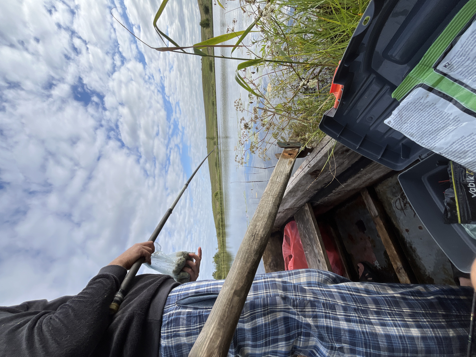 The image captures a person standing in a wooden boat on a lake, holding a fishing rod and a plastic bag containing fishing bait. The person is wearing a black sweatshirt and blue and white checkered pajama pants. The boat is partially filled with fishing gear, including a gray tackle box and a red plastic bag. The background shows a calm lake with green hills and a partly cloudy sky. The scene appears to be a serene fishing excursion in a natural setting. The location is identified as Dunilavichy Village Council, Belarus.