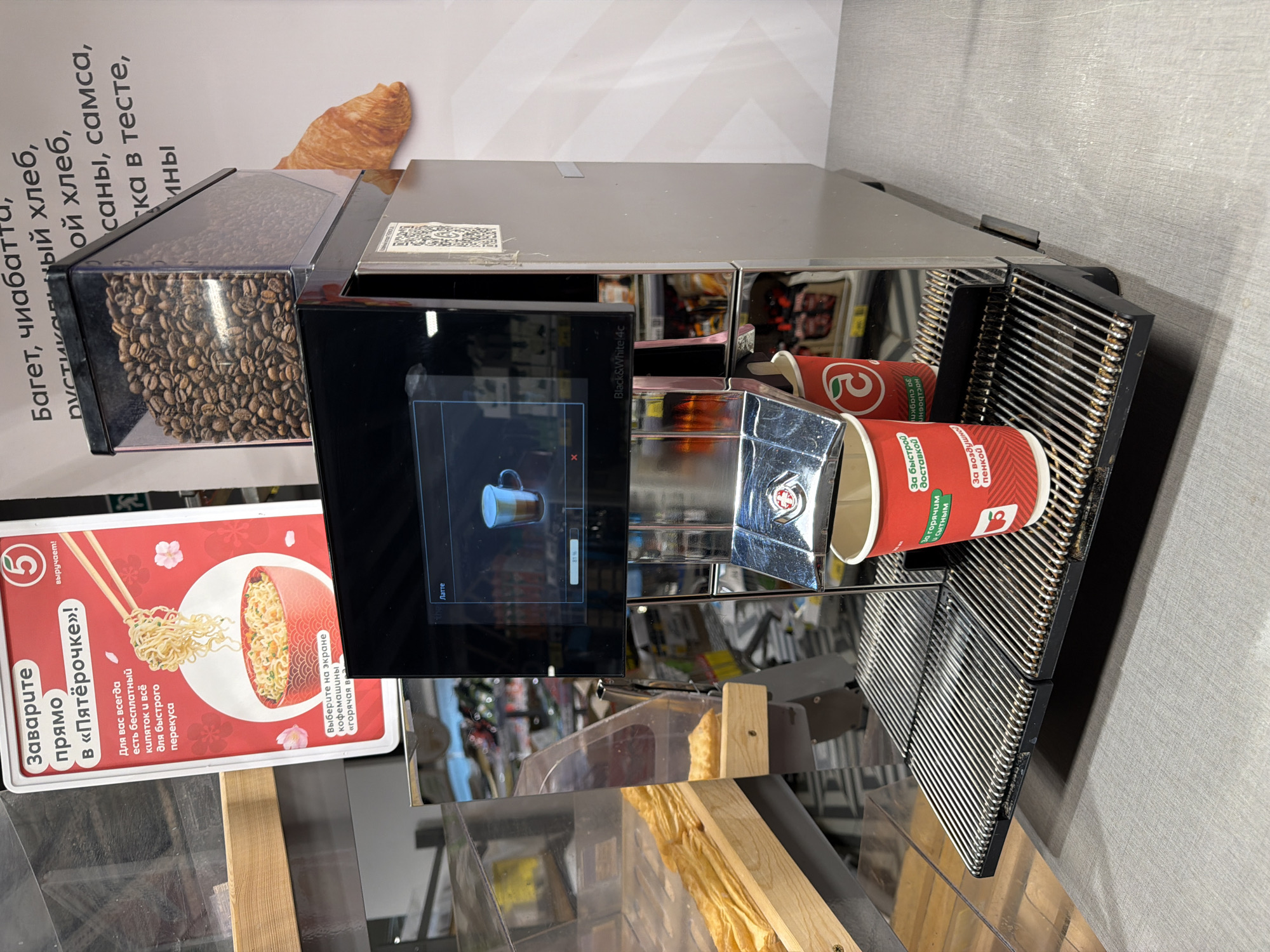 A close-up, eye-level, outdoor shot captures a commercial coffee machine in a retail setting. The machine, predominantly silver with black accents, features a digital display screen showcasing a "Latte" beverage in progress, indicated by an 81% completion bar. Above the screen, a clear plastic container holds roasted coffee beans. To the right of the coffee machine, a beige croissant is visible against a white poster. The machine's front includes a stainless steel drip tray with a red paper cup positioned on it, awaiting coffee. Behind the machine, shelves stocked with various food items are dimly visible. To the left, a display of pastries and baked goods is evident. The scene is illuminated by artificial lighting. The poster on the wall includes Cyrillic text, suggesting the location is in Russia. A red sign with a bowl of noodles and chopsticks, also in Cyrillic, is prominently displayed on the left. This specific setting is identifiable as the interior of a "Pyaterochka" supermarket branch in Semiluki, Russia, based on the signage.