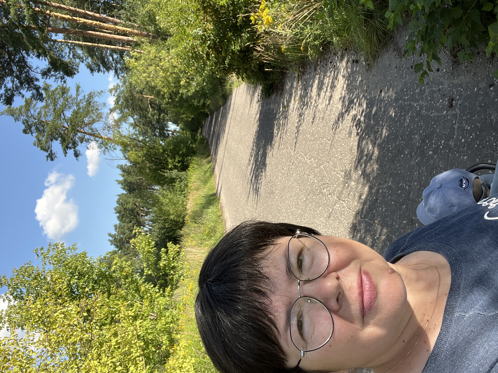 The image is a selfie taken from a low angle on a sunny day in a park. A woman with short black hair and glasses is in the foreground, looking at the camera. She is wearing a dark blue shirt and a thin gold chain necklace. Behind her, a paved path stretches into the distance, lined with green trees and foliage. The path is dappled with sunlight and shadows. A light blue baseball cap with a cartoon face on it is visible at the bottom right of the frame, suggesting the woman may be with a child or carrying something. The overall scene depicts an outdoor setting in a natural environment. The location of Balashikha, Russia, is not discernible from the visual information in the image.