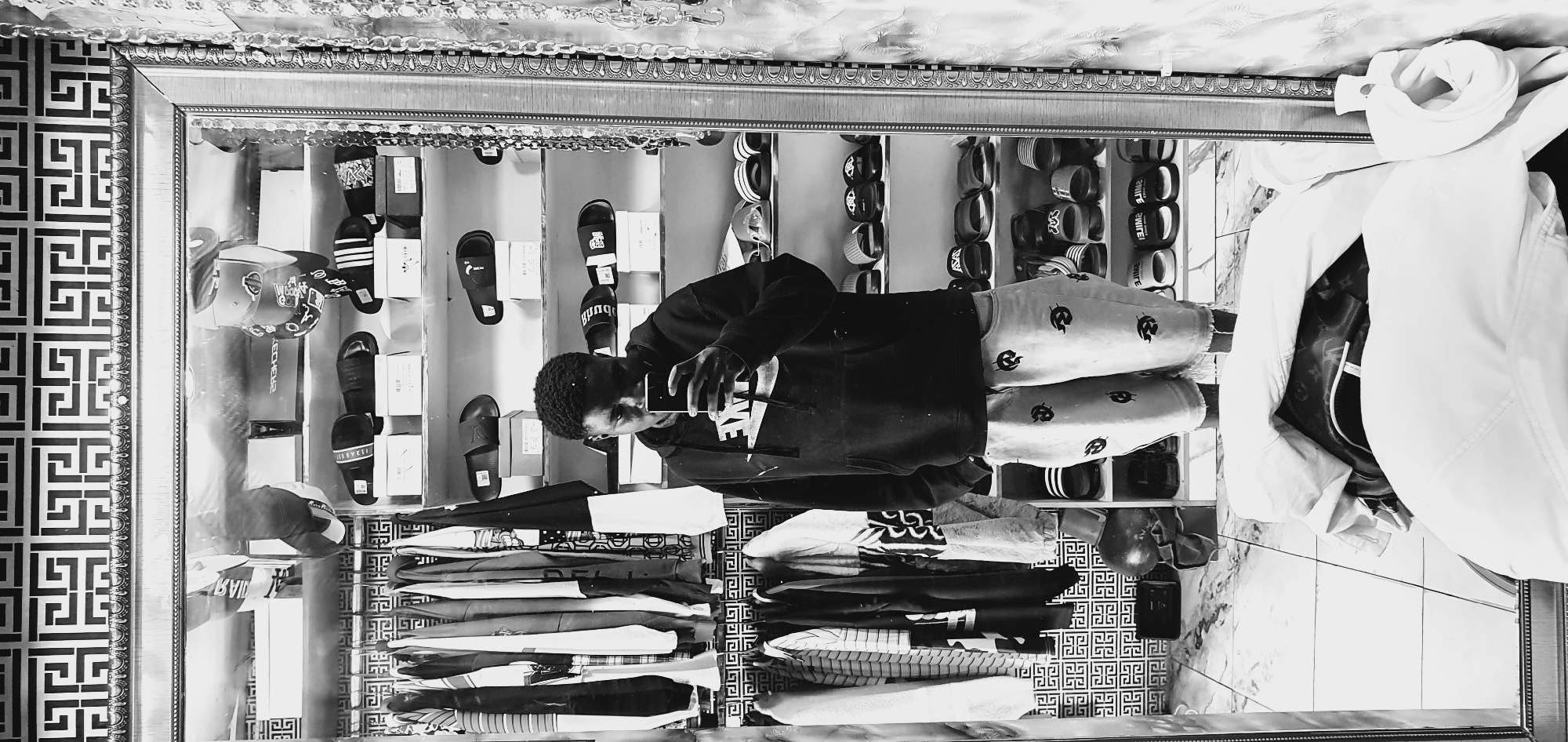 A full-body, black and white, mirror selfie captures a young man in Bukuru, Nigeria. He stands in what appears to be a store or a large closet, with clothing racks to his left and shelves of footwear behind him. The man, wearing a Nike hoodie and patterned shorts, holds a phone up to take the photo. The background is decorated with a geometric pattern. The floor is tiled with a marble-like pattern. The overall scene suggests a retail environment or a personal collection of fashion items.