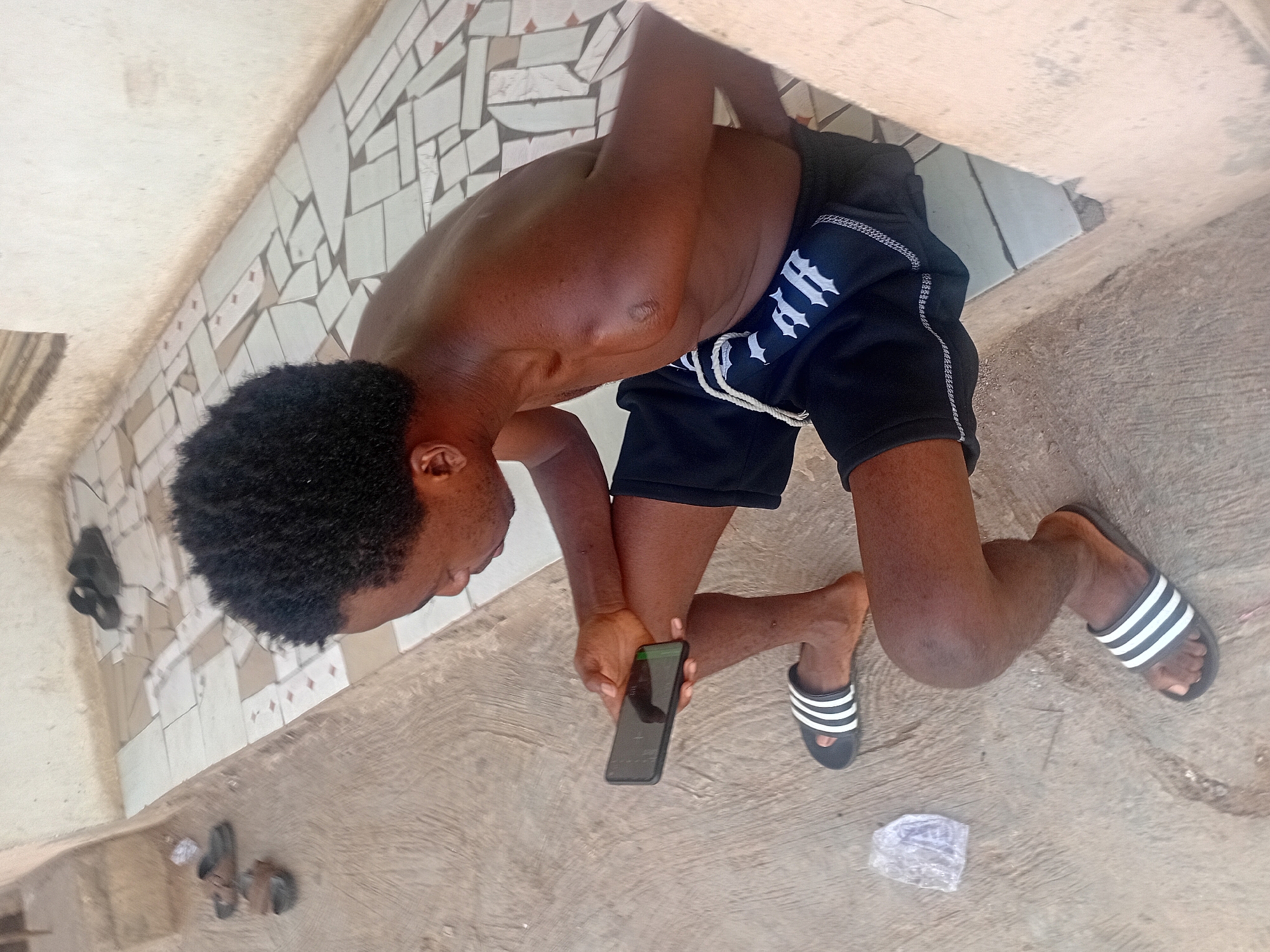 A dark-skinned male is depicted kneeling on a concrete surface, leaning forward and looking down intently at a black smartphone held in both hands. He has short, curly black hair and is shirtless, wearing black athletic shorts with white drawstrings and a white gothic-style logo, possibly reading "RTII" or similar, on the left thigh. On his feet, he wears black slides with three prominent white stripes, characteristic of the Adidas brand.
The immediate setting is a casual outdoor or semi-enclosed area. The floor is predominantly concrete, but along the edge of the frame, there is a low wall or raised platform covered with small, irregularly shaped light-colored tiles. Further back on this tiled surface, two other pairs of sandals are visible. A small, crumpled piece of clear plastic or paper lies on the concrete near the person's right foot. The lighting suggests it is daytime. The smartphone screen displays green elements at the top, indicating an active interface. The person appears focused on the device.