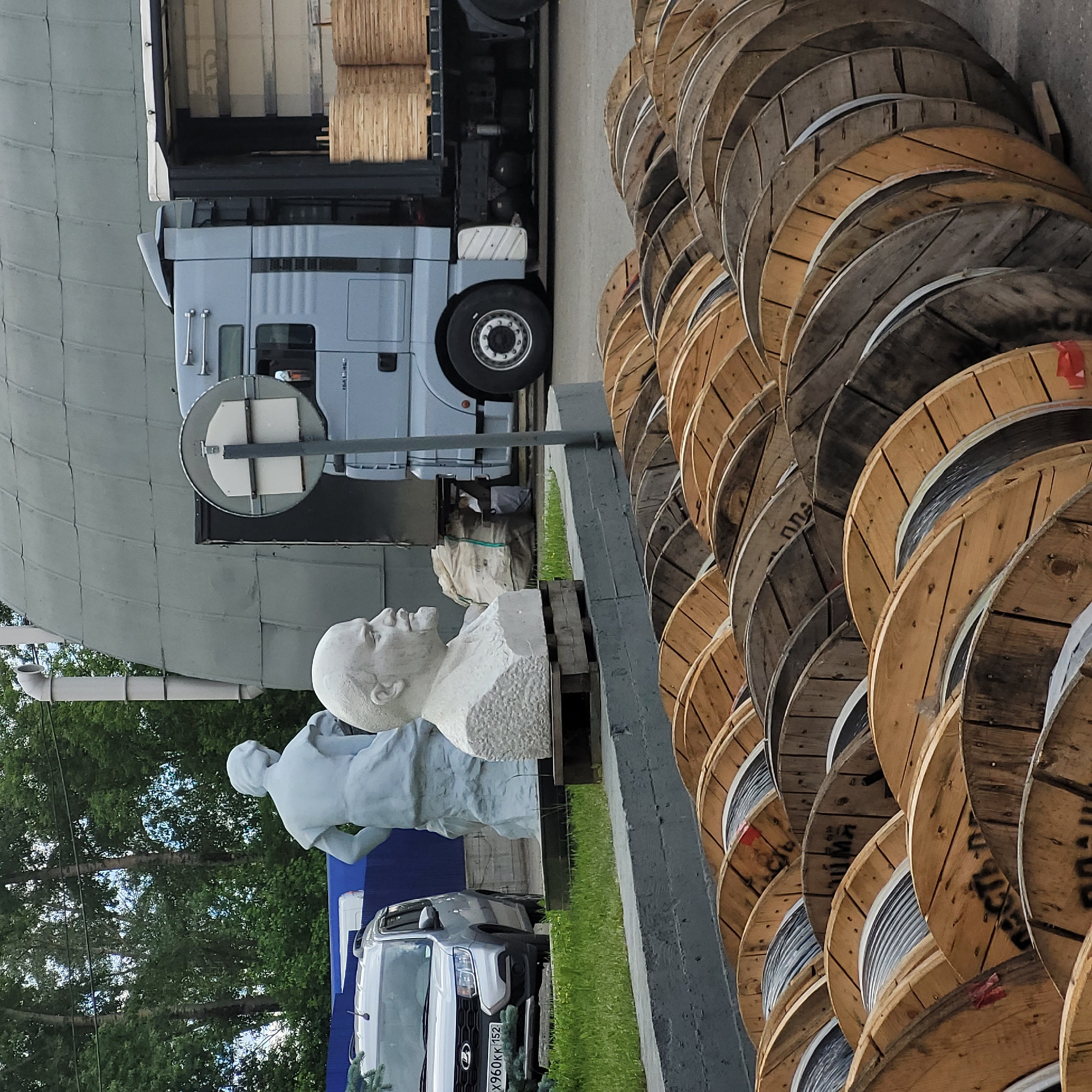 The image captures an outdoor industrial or storage area featuring a parked semi-truck, a row of large wooden cable spools, and two statues.
**Subjects:**
A grey MAN semi-truck with an open trailer is parked on the left side of the frame, seemingly undergoing loading or unloading, as its trailer contains wooden spools. Adjacent to the truck, a white bust of Vladimir Lenin and a grey full-body statue of a figure (possibly a worker or revolutionary) are displayed on a raised platform. To the right, a long line of numerous large, stacked wooden spools, likely for industrial cables, stretches into the distance. A light-colored car is partially visible behind the statues. In the background, there is a large, grey, curved building structure and green trees.
**Activity or event:**
The scene depicts a static moment in an industrial setting. The truck is stationary, and the cable spools are stored in an organized manner. The statues appear to be a permanent fixture in this outdoor space.
**Notable details:**
The scene takes place during the daytime, under what appears to be an overcast sky, providing soft, diffused lighting. Green grass is visible between the paved areas and the statues. The cable spools show signs of wear and varying ages. The car, a Lada Granta or similar model, bears a Russian license plate with the code "X960KK 152," where "152" designates the Nizhny Novgorod Oblast in Russia.
**Visible text:**
Text on the car's license plate reads "X960KK 152." On some of the wooden cable spools, faint Cyrillic lettering like "КАБЕЛЬ" (KABEL, meaning "cable") and other numbers are visible. The brand "MAN" is visible on the truck's grille.