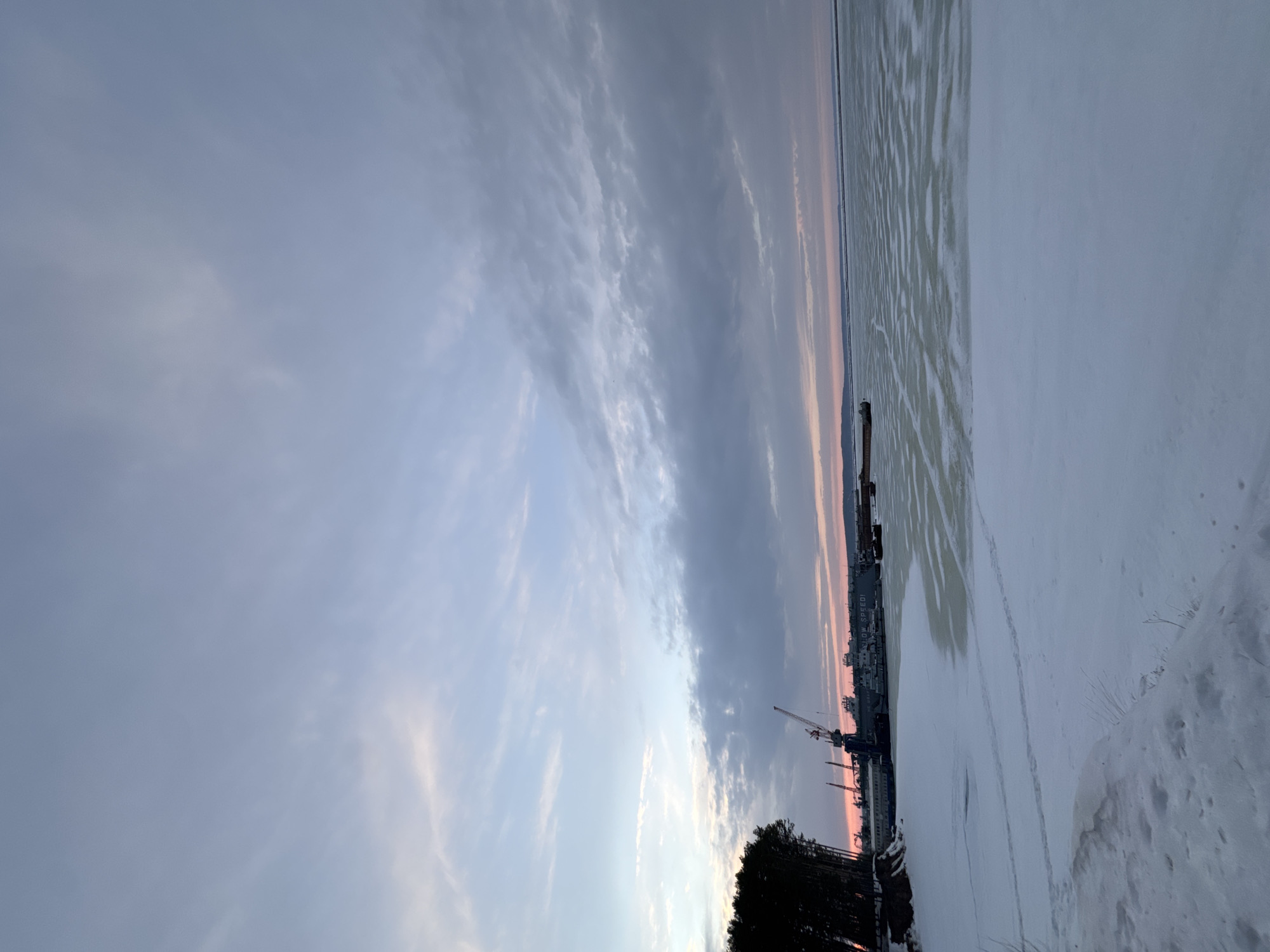 The image shows a winter scene in what appears to be the city of Chaikovsky, Russia. The sky is dominated by clouds, transitioning from lighter hues near the horizon to darker shades above. The ground is covered in snow, and the edge of a body of water, likely the Kama River, is visible, with ice formations present. A harbor or industrial area is situated along the shoreline, featuring cranes and possibly ships or docked vessels. A line of trees is at the edge. The general environment seems cold, bleak, and somewhat industrial. The angle is vertical.