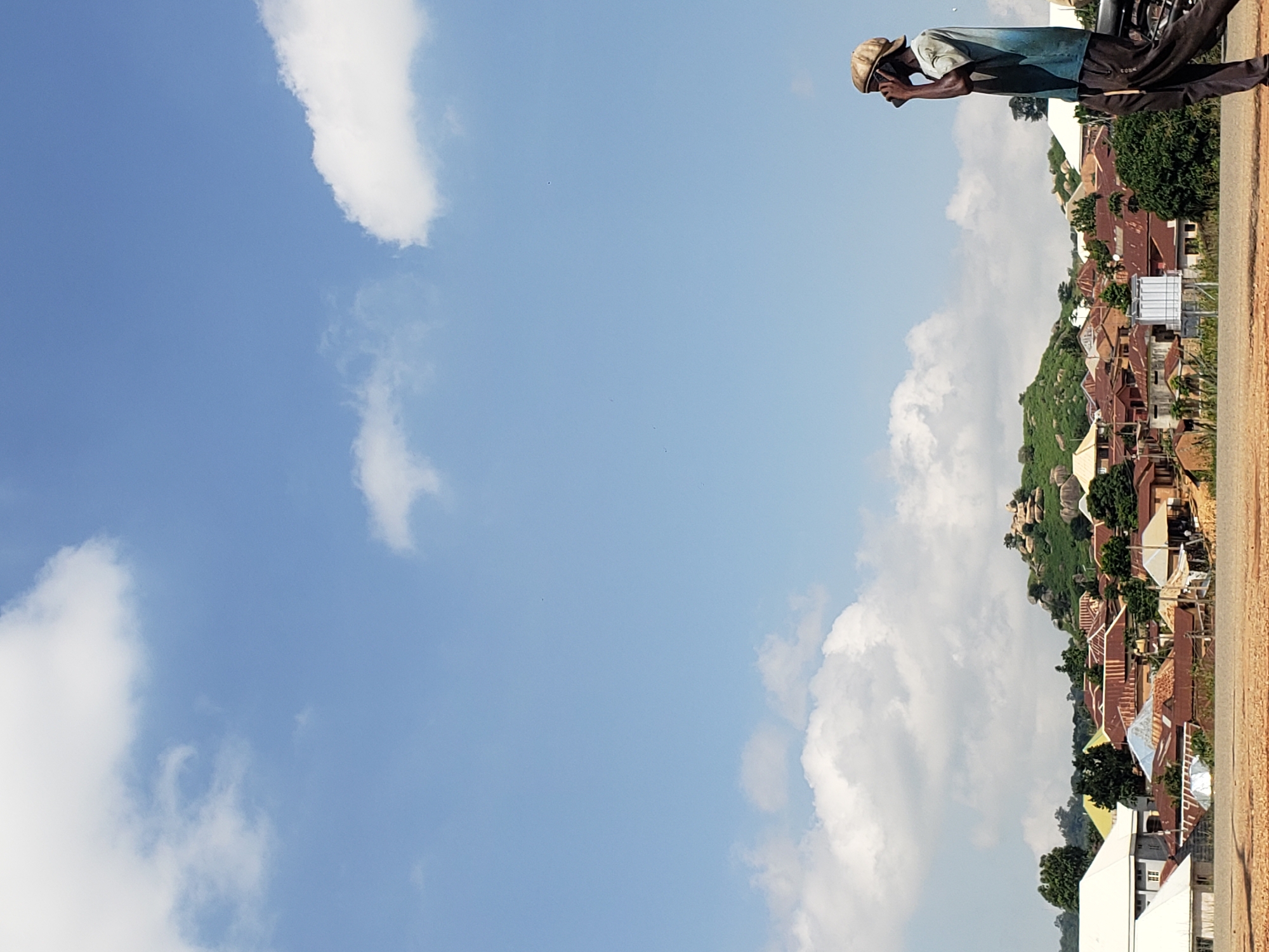 A man in a faded blue t-shirt and dark pants walks on a dirt path, talking on a cell phone. He wears a light brown cap. Behind him, a village with many houses with corrugated metal roofs stretches across a green, hilly landscape. The sky is bright blue with scattered white clouds. The image appears to be taken in the city of Jos, Nigeria, based on the architectural style of the buildings and the vegetation.