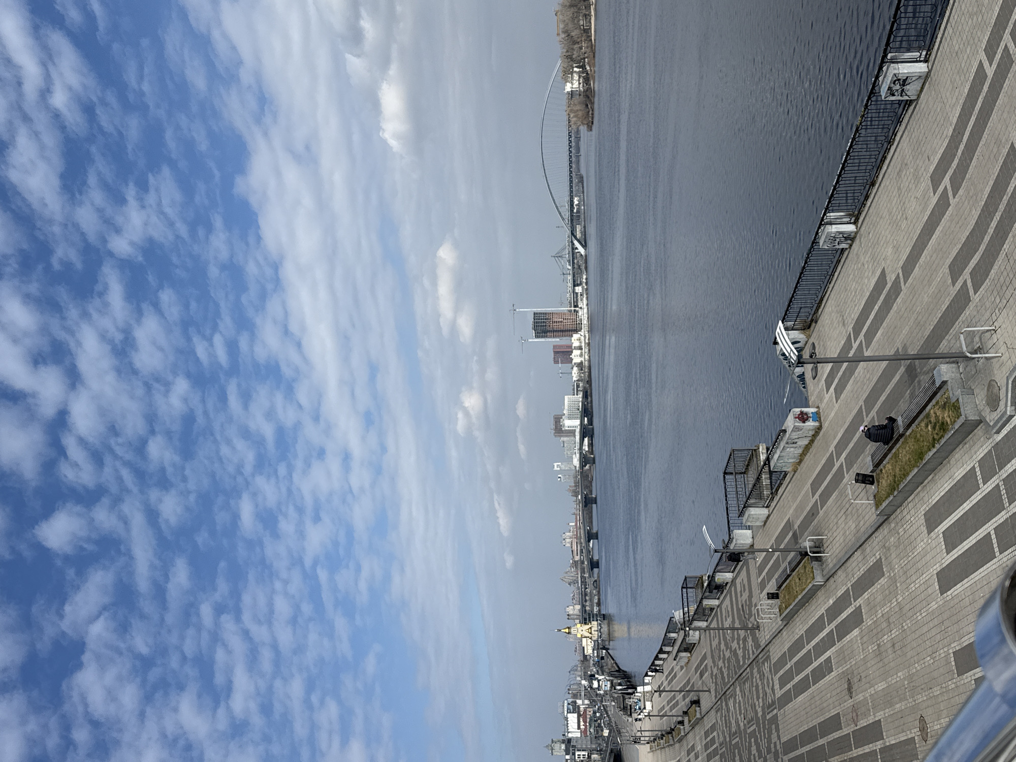 The image depicts a panoramic view of the Dnieper River in Kyiv, Ukraine, observed from an elevated position on the Podil Embankment, looking southeast.
The foreground features a wide, paved pedestrian promenade constructed from light and dark grey rectangular paving stones. The paving includes rectangular beds with sparse grass and grey metal benches without occupants, except for one instance. A single individual, dressed in dark clothing with a light-colored hat, is seated on a bench in the lower right quadrant, facing away from the viewer towards the river. Multiple grey metal lampposts and white metal bike racks are spaced along the promenade. A dark metal railing with concrete supports borders the embankment edge, separating it from the river; graffiti is visible on some concrete sections. A chrome-colored railing, part of the vantage point, is visible in the bottom left.
The Dnieper River, exhibiting a dark grey-blue hue with minor surface ripples, occupies the midground. Adjacent to the left bank, a yellow-domed, white-walled church (St. Nicholas Church on the Water) stands on a platform within the river. Further downriver, multiple bridges span the Dnieper. These include a multi-span concrete bridge, the distinct white arched Parkovy Pedestrian Bridge, and, further to the right, the large, incomplete arched structure of the Podilskyi Bridge, marked by prominent construction cranes and a tall, unfinished high-rise building with exposed rebar on the left bank.
The background comprises the Kyiv city skyline with various urban structures. The sky is blue with numerous scattered white clouds (altocumulus or stratocumulus). The right bank of the river features an undeveloped land strip with scattered trees.