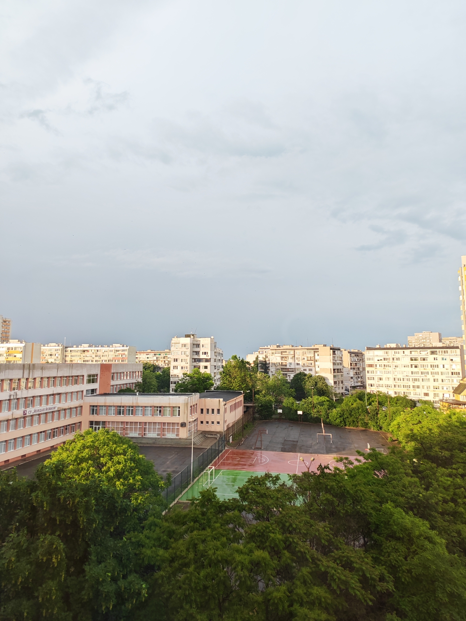 The image depicts an elevated view of a cityscape, likely in Burgas, Bulgaria, judging by the architectural style and Cyrillic text on a building. The sky is overcast with grey clouds. Several multi-story buildings, predominantly residential, dominate the middle ground. These buildings are light-colored with repetitive window patterns. In the foreground, there's a school building with a sports complex. The school building has a pink facade with horizontal rows of windows. The sports complex consists of basketball and soccer courts, partially obscured by lush green trees. The sports court is partially wet, possibly from rain. The scene is still and seemingly devoid of human activity.
