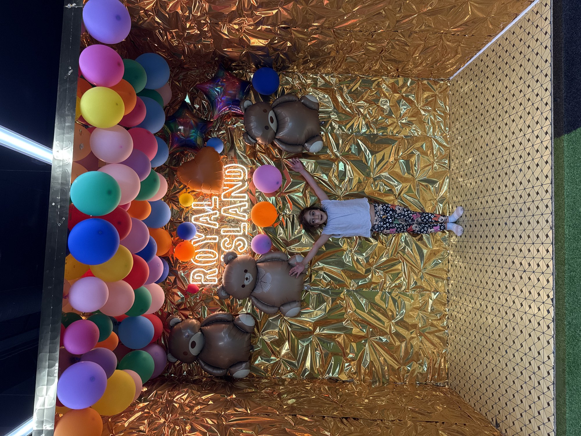 A young person lies on their back in a room decorated with shiny gold foil. They are wearing a white t-shirt and patterned leggings. The room is filled with colorful balloons, some clustered on the left side of the frame and others scattered around the person and the gold foil. Several large teddy bear-shaped balloons are also present. To the left of the teddy bears, a neon sign glows, spelling out "ROYAL ISLAND". The gold foil covers the walls and ceiling of the room, creating a reflective and festive atmosphere. The lighting suggests it is daytime, and the overall mood is playful and celebratory. The scene appears to be an indoor event or party space, possibly in Sochi, Russia, given the location context.