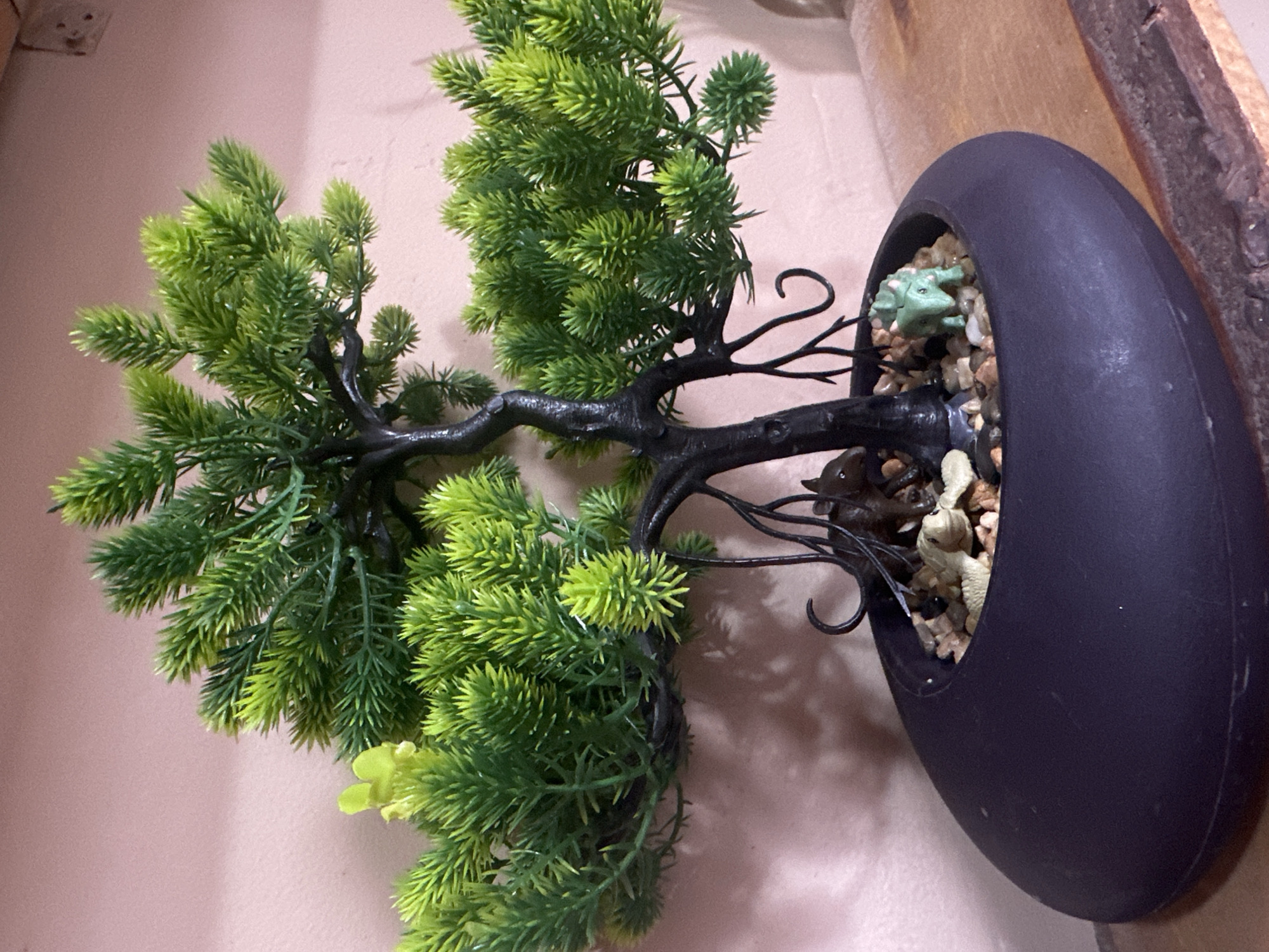 An artificial bonsai tree with a dark trunk and green foliage sits in a black oval pot filled with small stones. Tiny toy animals, including what appears to be a dinosaur and other small creatures, are nestled among the stones at the base of the tree. The pot is placed on a wooden surface against a pinkish-toned wall. The scene is indoors, and no specific location in Tashkent, Uzbekistan, can be identified from the image.