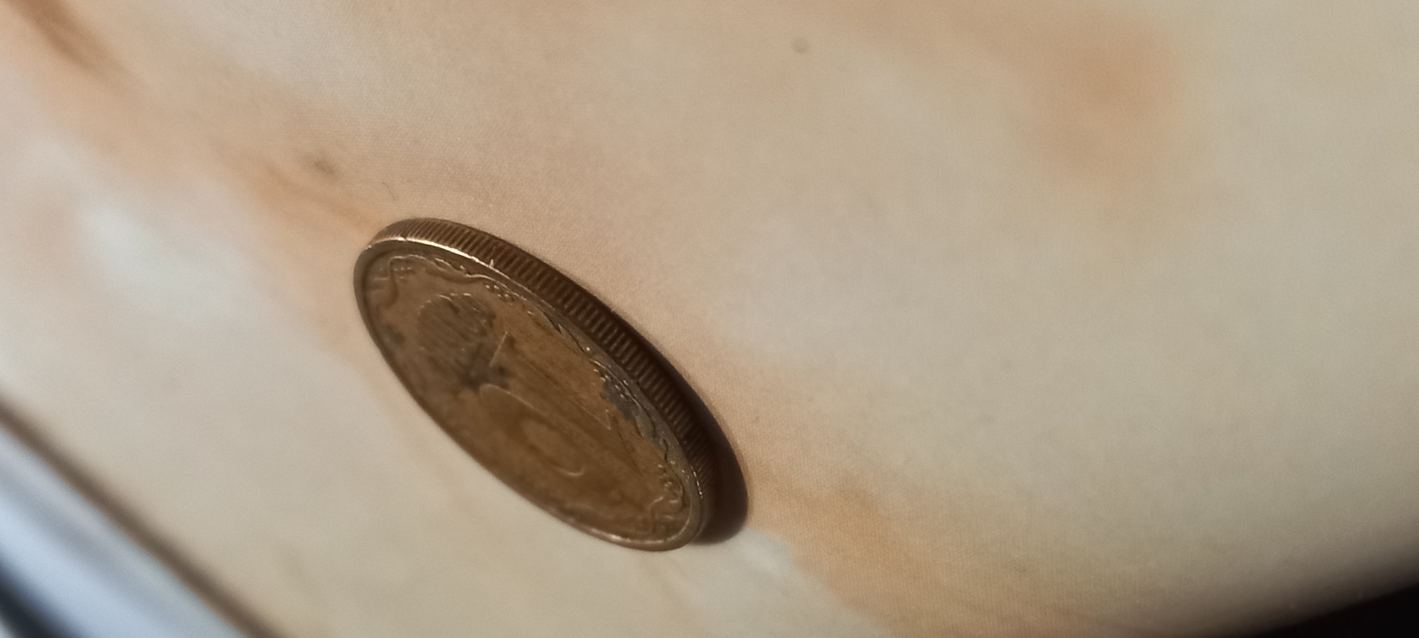 The media file presents a close-up, angled photograph of a single coin resting on a light-colored, discolored surface. The coin, likely metallic with an aged golden-brown patina, features a distinctly reeded edge. Its obverse side is partially visible, showing raised design elements including the number "2" and what appears to be part of the Ukrainian national emblem (tryzub), identifying it as a Ukrainian 2 Hryvnia coin.
The coin is positioned diagonally, with a subtle shadow cast beneath it, indicating a light source from above. The surface beneath is pale, possibly beige, and exhibits several irregular, brownish stains or areas of discoloration, particularly concentrated around the coin and in the upper background, suggesting age or wear. The texture of this surface appears soft, potentially fabric or aged paper. In the bottom-left corner of the main image, a blurry, dark, curved object is partially visible, but unidentifiable.
The scene depicts a still life; no specific activity or event is taking place. The setting appears to be indoors, so the time of day and weather are not discernible. The presence of a Ukrainian coin aligns with the provided location context of Kryvyi Rih, Ukraine. No other explicit text is clearly visible on the coin or the surrounding elements.