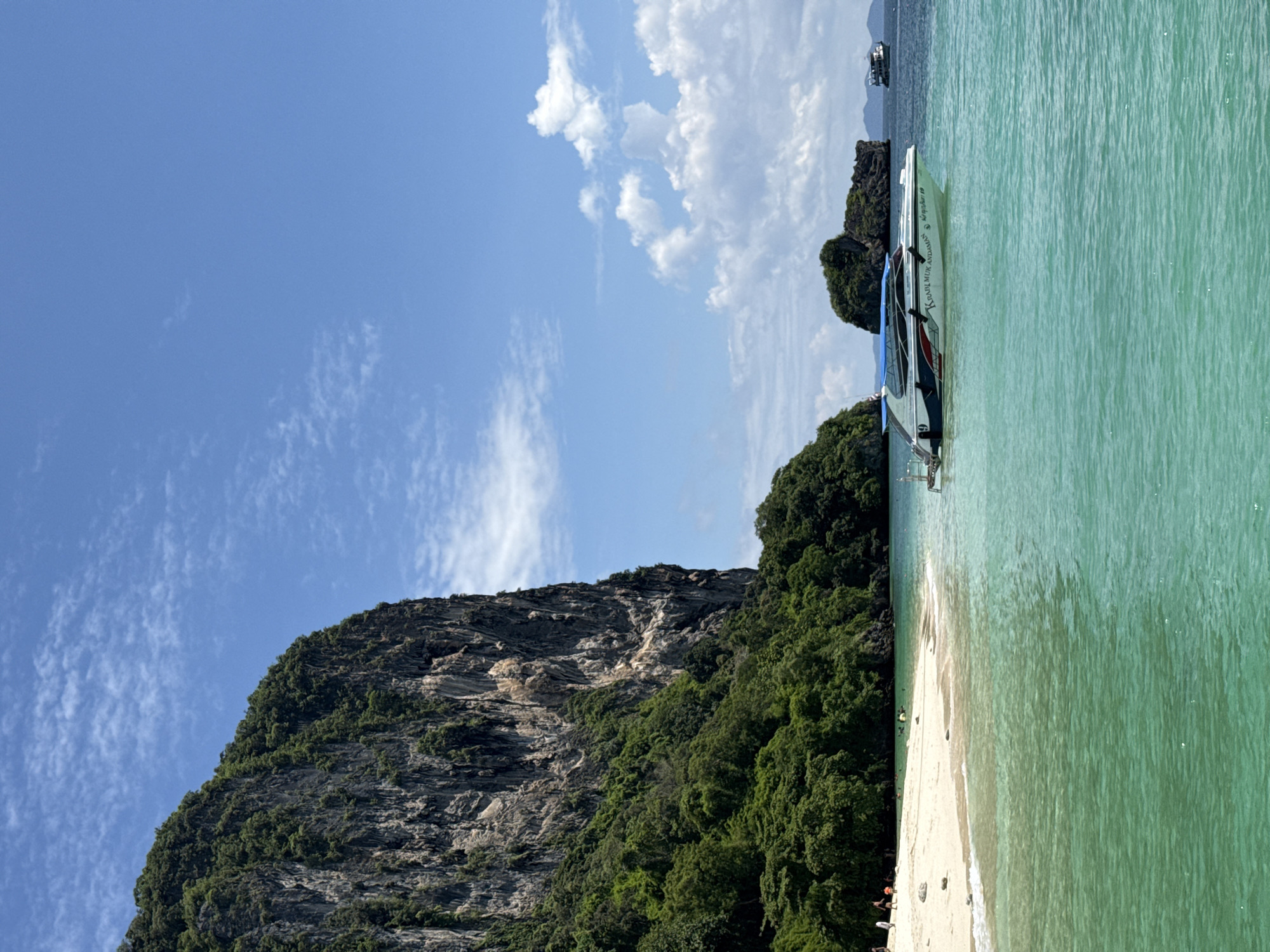 In Ao Nang, Thailand, a serene beach scene unfolds with a dramatic limestone karst rising on the left. The cliff face, covered in lush green foliage, dominates the background. A white sandy beach stretches along the coastline, with a few distant figures visible enjoying the sun and sea. The turquoise waters of the Andaman Sea are clear and inviting, gently lapping at the shore. In the foreground, a sleek speedboat, emblazoned with "KRABI MUK ANDAMAN," cuts through the water, its blue canopy providing shade. Another boat can be seen in the distance. The sky above is a vibrant blue, dotted with fluffy white clouds, suggesting a bright and sunny day. The overall impression is one of tropical paradise, with natural beauty and recreational activities coexisting harmoniously.