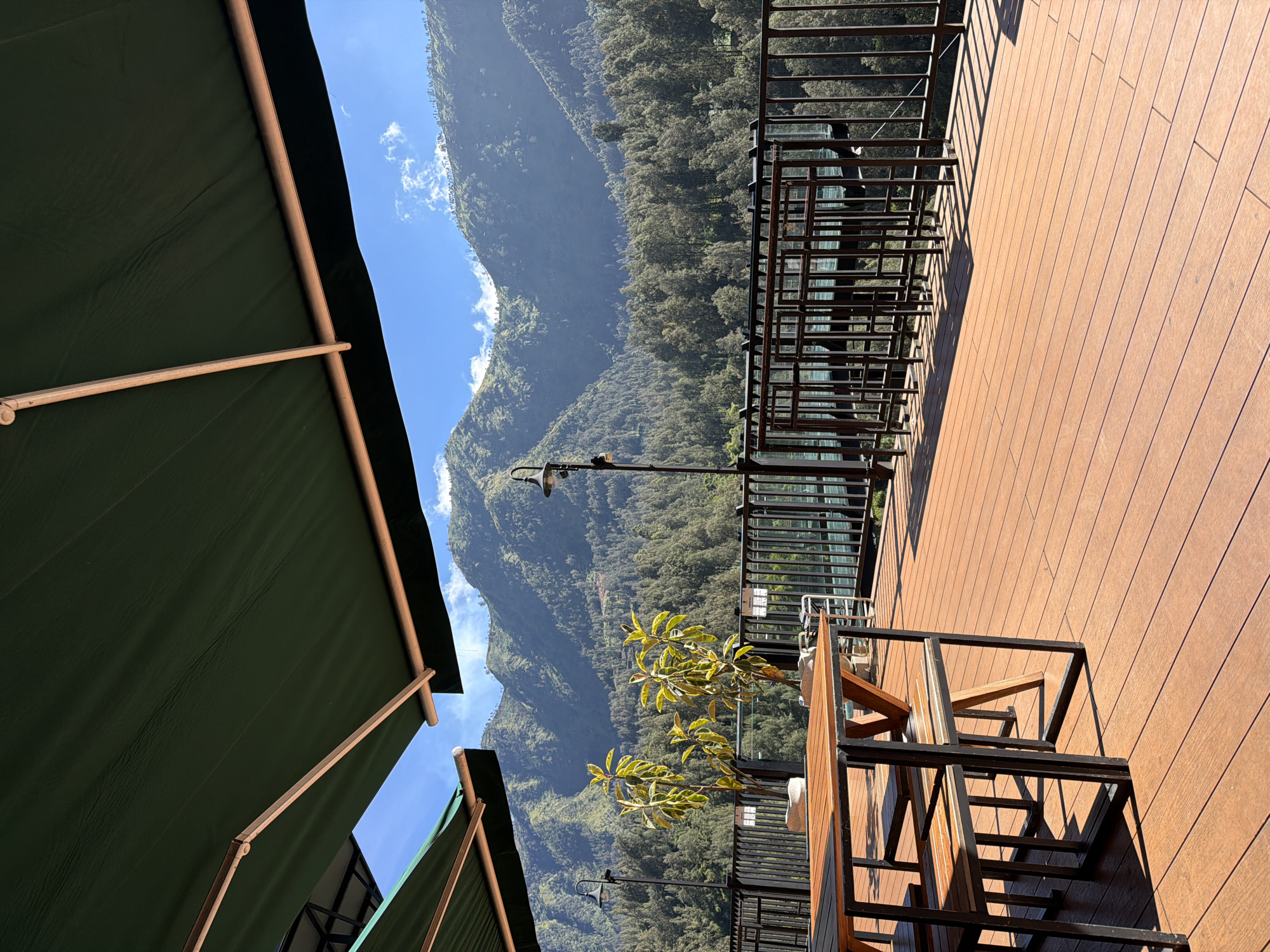 The image depicts an outdoor seating area with a wooden deck and metal tables and chairs, overlooking a lush, forested mountain range under a bright blue sky with scattered clouds. Two green awnings are visible overhead, casting shadows on the deck. In the foreground, a wooden table with a black metal frame is set with chairs. A small tree with green leaves stands to the left of the seating area, near a lamp post. A black metal railing separates the deck from the forested slope below. The scene appears to be a tranquil, natural setting, likely a restaurant or cafe with a scenic view. The location is identified as Jetak, Indonesia.