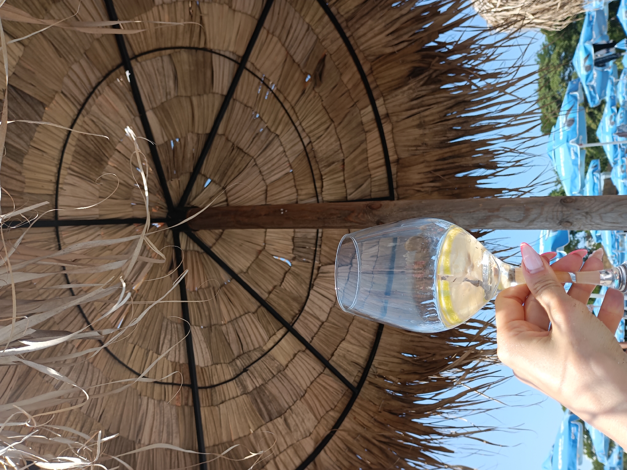 A person's hand with pink-tipped fingernails holds a wine glass filled with a pale yellow liquid and lemon slices under a thatched umbrella at the beach. The umbrella's natural fiber material is visible, along with its structural supports. In the background, a clear blue sky is visible, along with rows of blue beach umbrellas and green trees. The location is likely the beach in Burgas, Bulgaria.